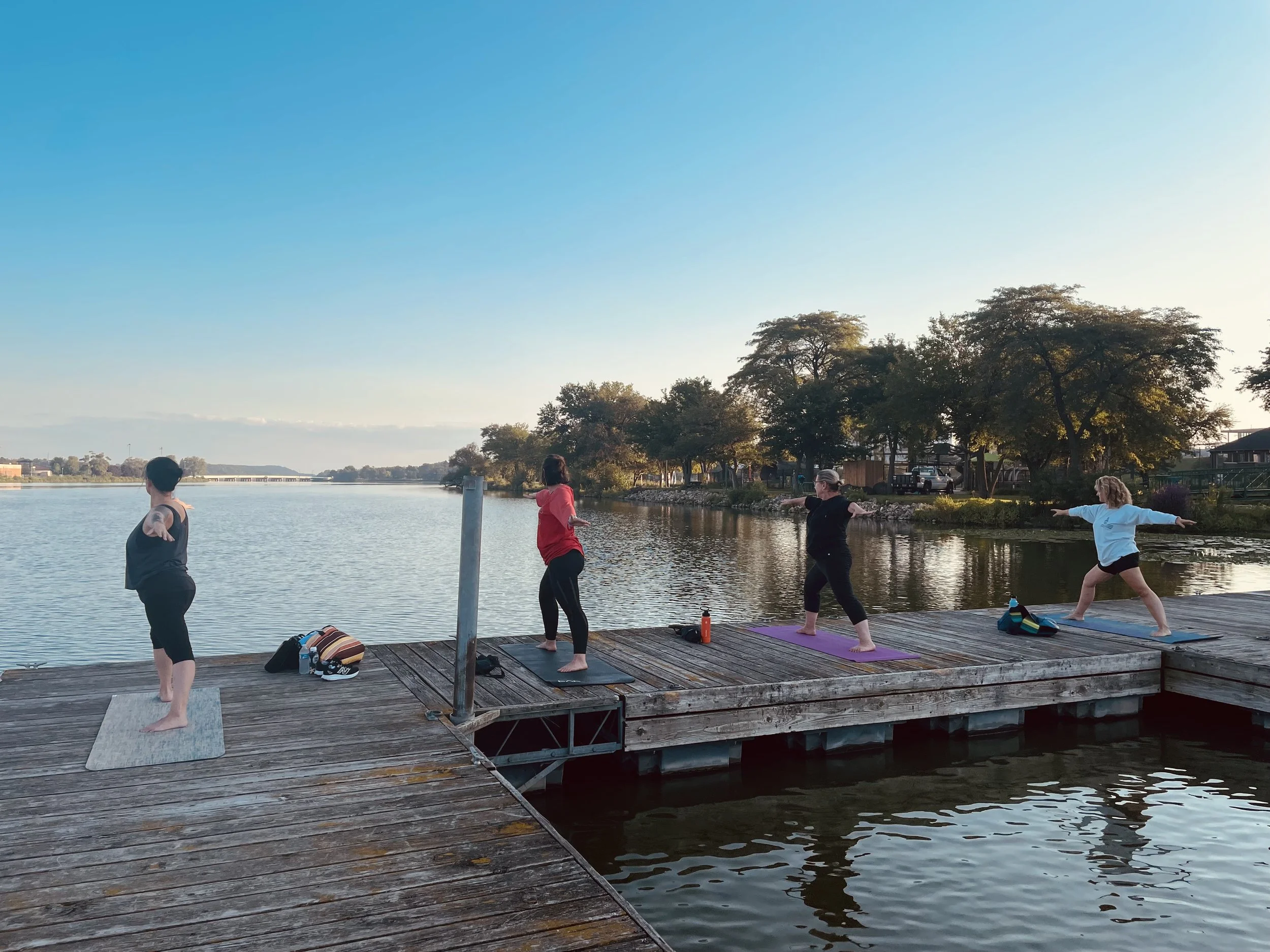 Four women practicing yoga on a dock beside a river at sunset, with trees and houses in the background.