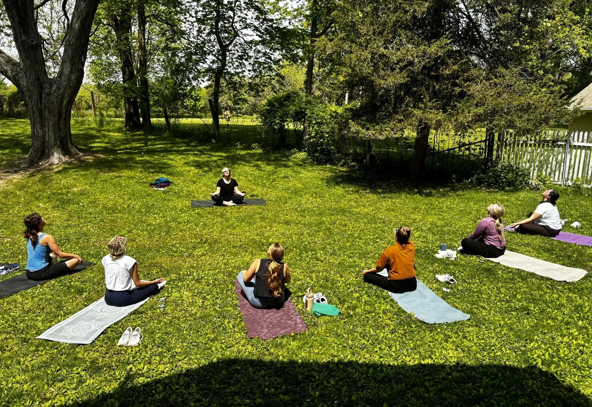 A group of people practicing yoga outdoors on mats in a grassy yard surrounded by trees and a white fence on a sunny day.