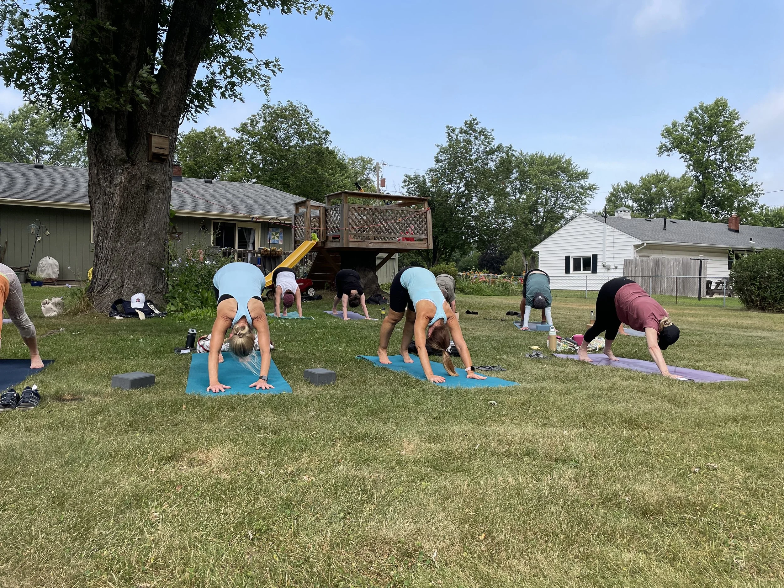 People practicing yoga outdoors in a backyard on yoga mats, with trees, houses, and a clear blue sky in the background.
