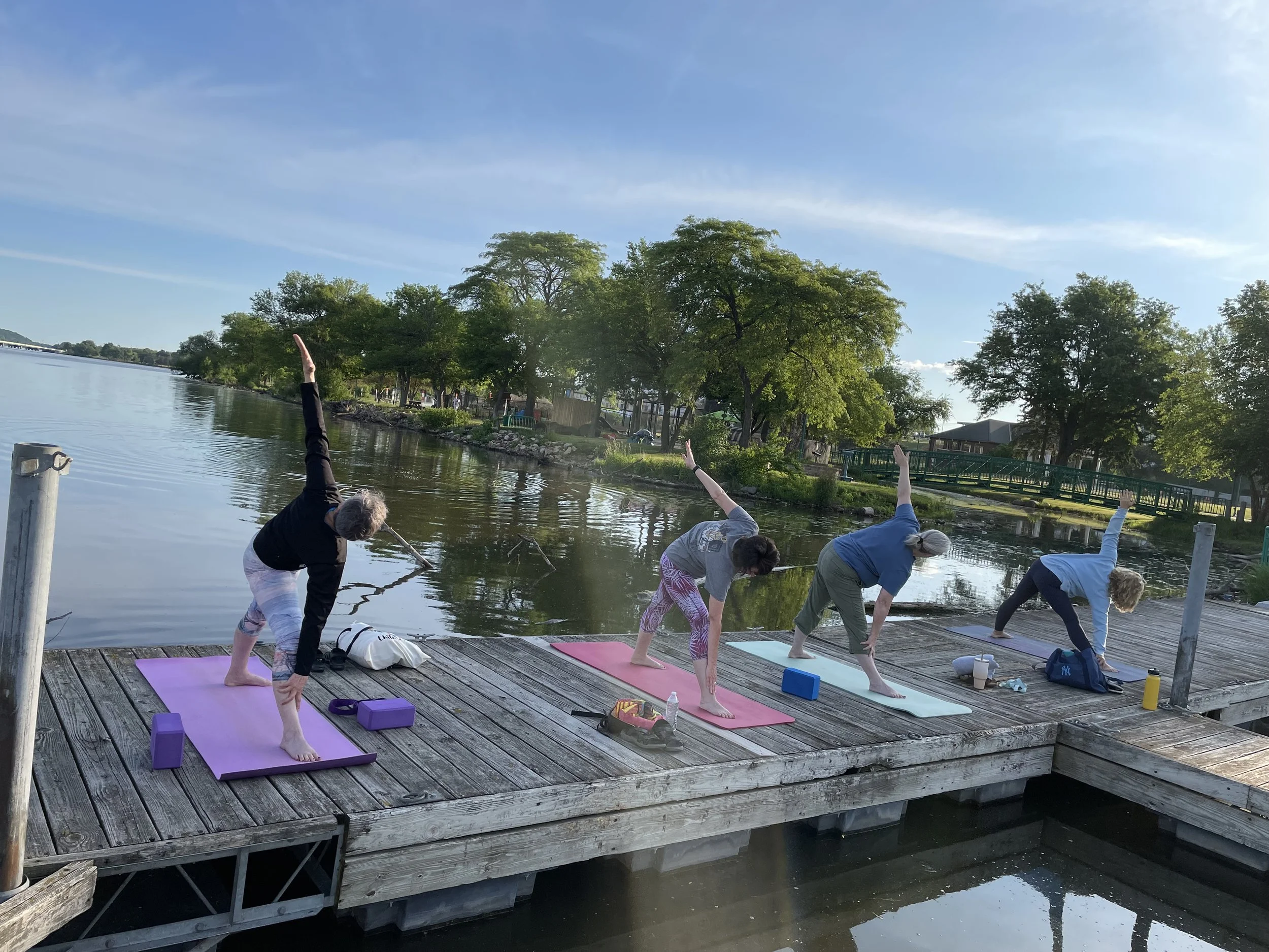 People practicing yoga outdoors on a wooden dock by a lake during the daytime. They are on colorful yoga mats near water, with trees and a clear sky in the background.