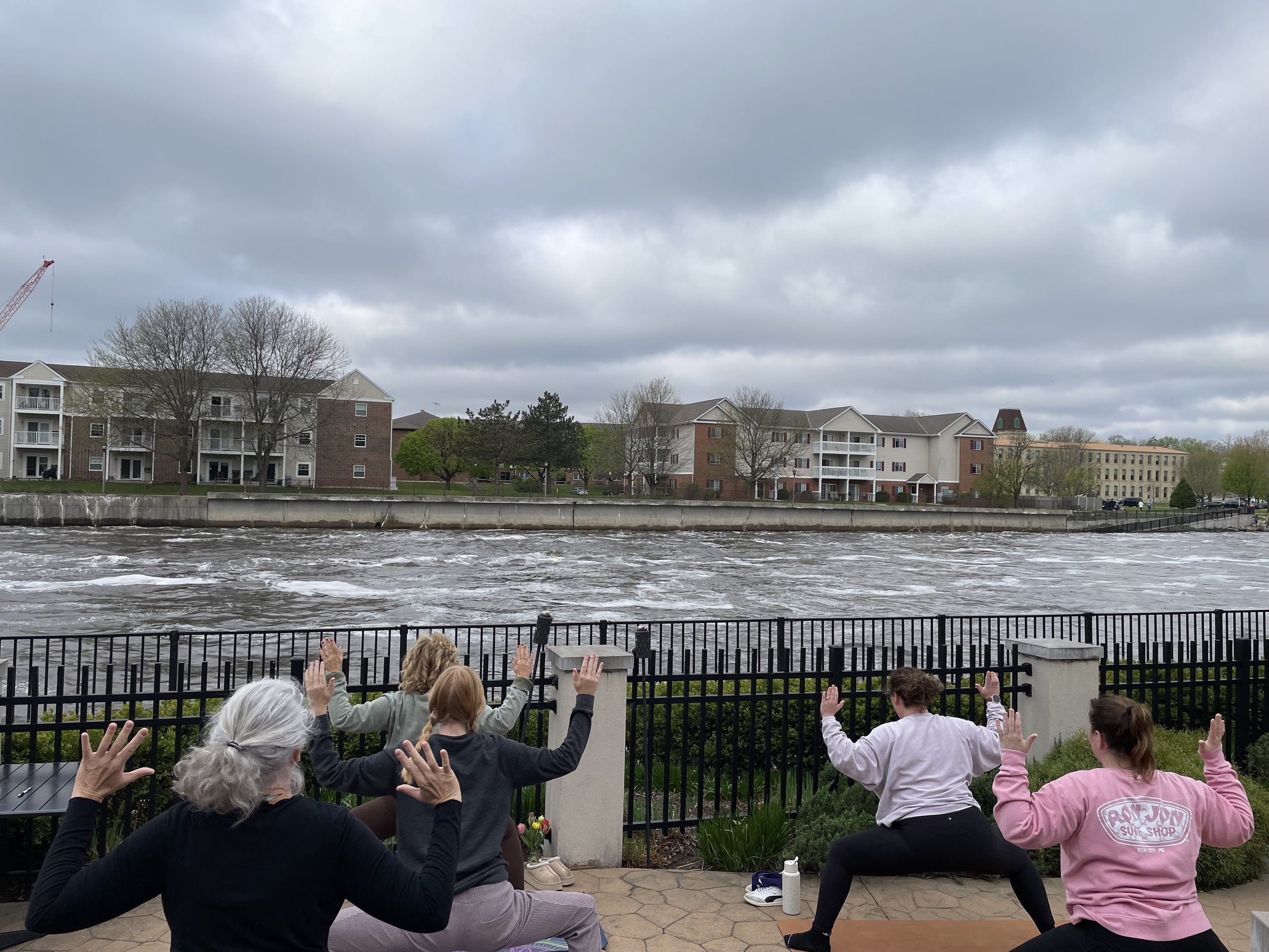 Group of elderly and young people doing yoga outdoors by a river, with apartment buildings and cloudy sky in the background.
