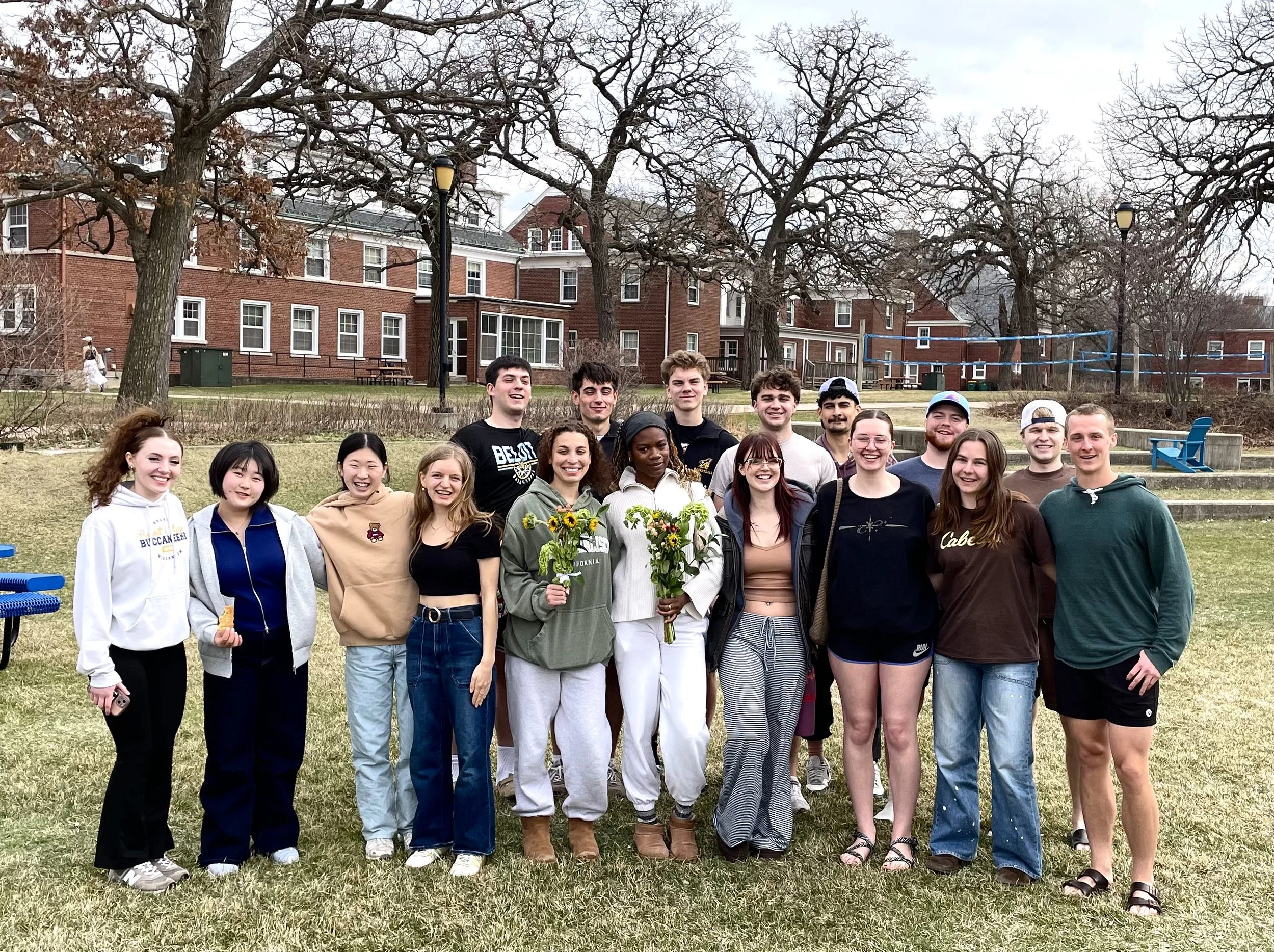 Group of diverse young adults smiling and standing outdoors on a grassy area, with some holding bouquets of flowers. Background includes trees, brick buildings, and a volleyball net.