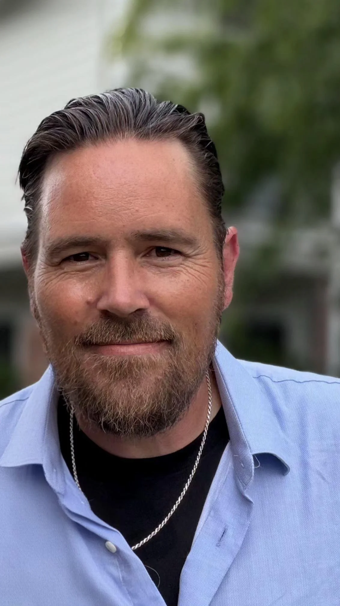A man with a beard, smiling, wearing a light blue shirt, black t-shirt, and silver chain, outdoors with blurred greenery in the background.