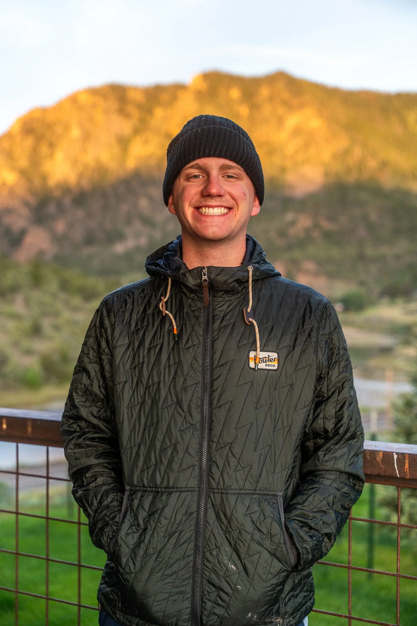 A young man smiling outdoors wearing a black beanie and black jacket, with a mountain and a partly cloudy sky in the background.