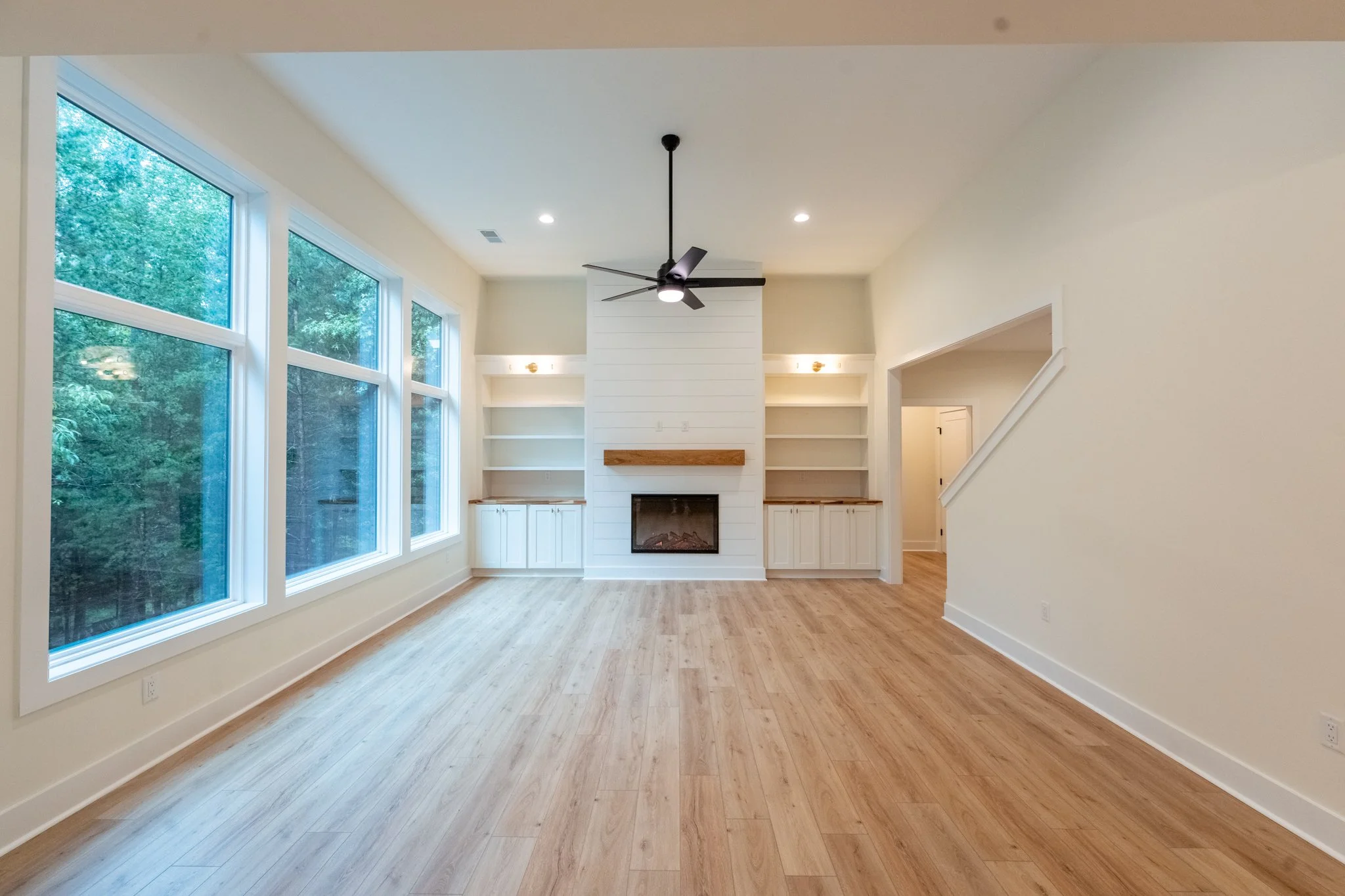 Empty living room with large windows, a fireplace, built-in shelves, and a ceiling fan.