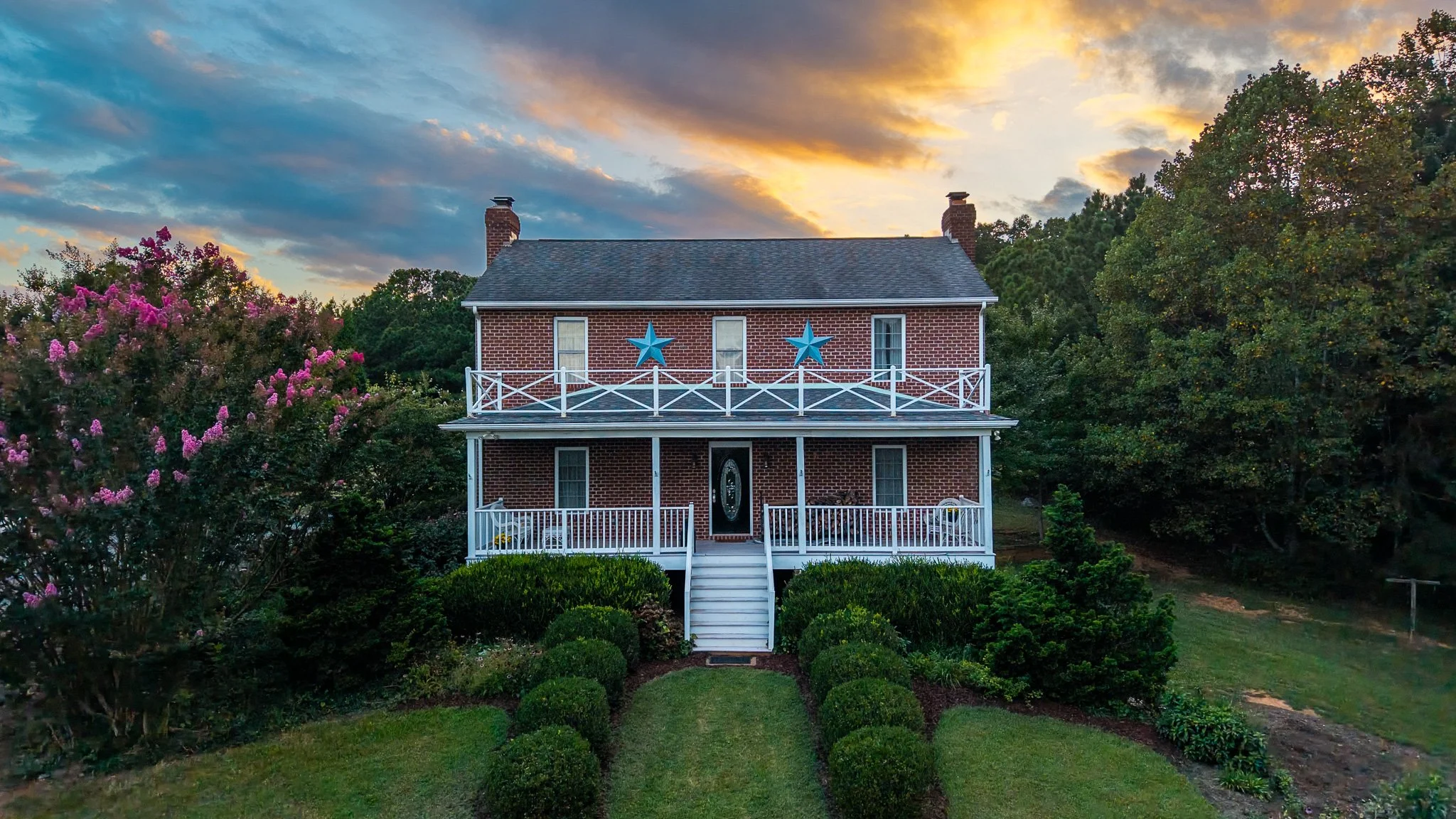 A brick house with two stories, a front porch, and two blue star decorations on the second-floor railing, surrounded by greenery and flowering bushes under a sunset sky.