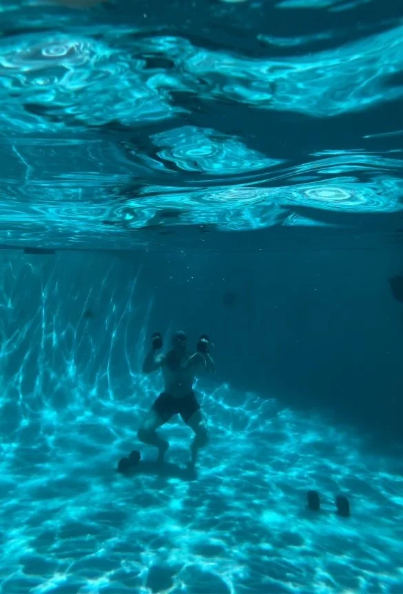 Person underwater in a swimming pool, mid-air, with arms raised and wearing swim shorts. Wellness Membership St. Louis | Breath & Exposure STL