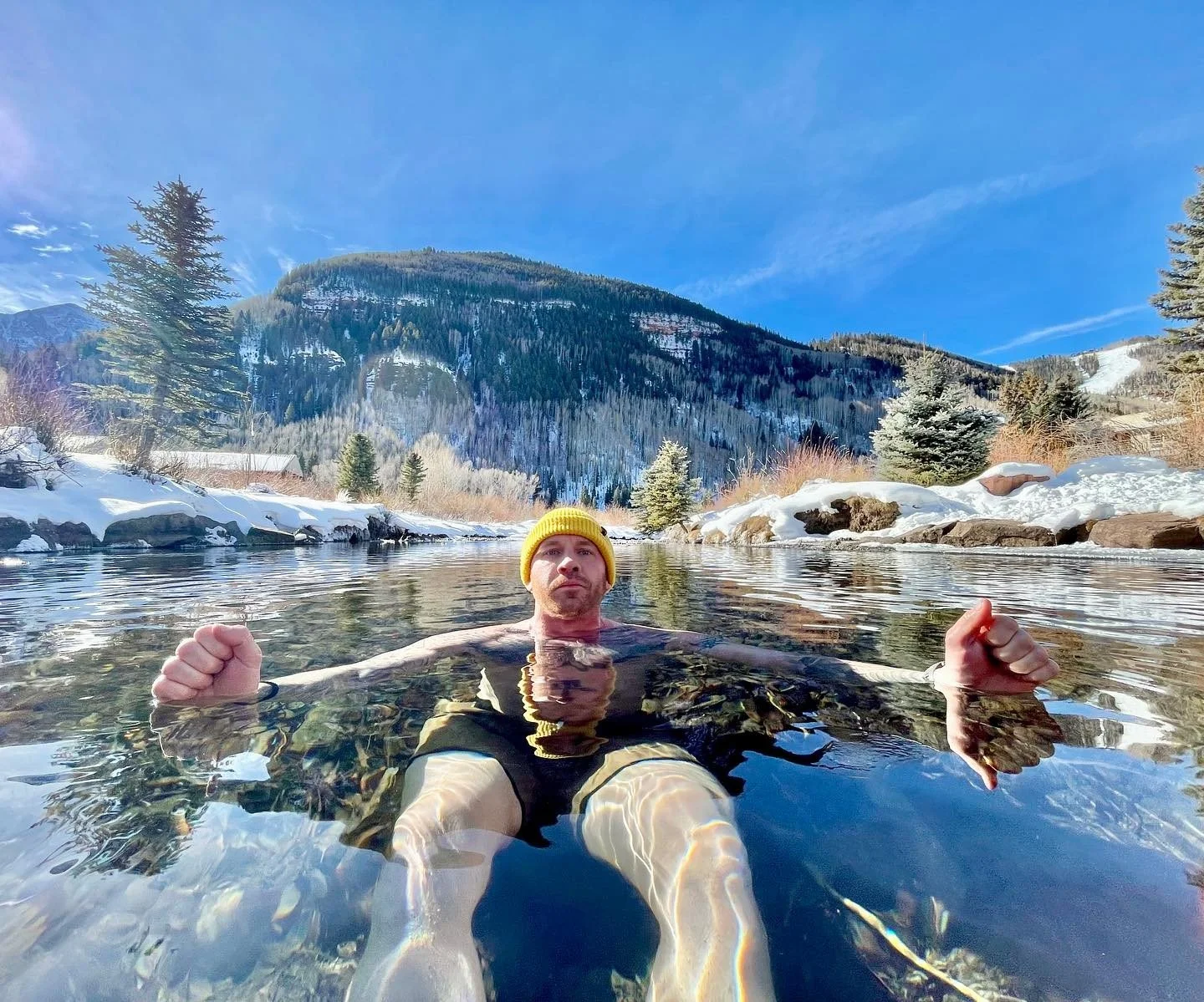 Man wearing a yellow beanie relaxing in a river with snow-covered banks in a mountainous winter landscape. Wellness Membership St. Louis | Breath & Exposure STL