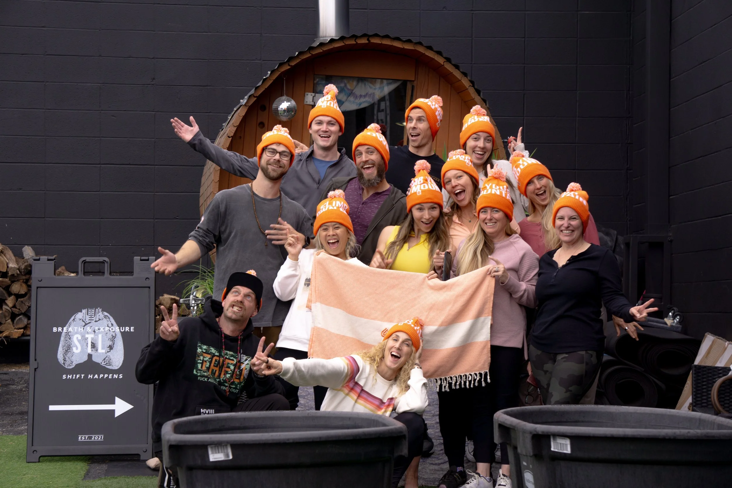 Group of people wearing orange knit hats with pom-poms, smiling and posing for a photo outside a small wooden structure, holding a pink and white towel, some making peace signs, against a black wall with a sign pointing right. Wellness Membership St.