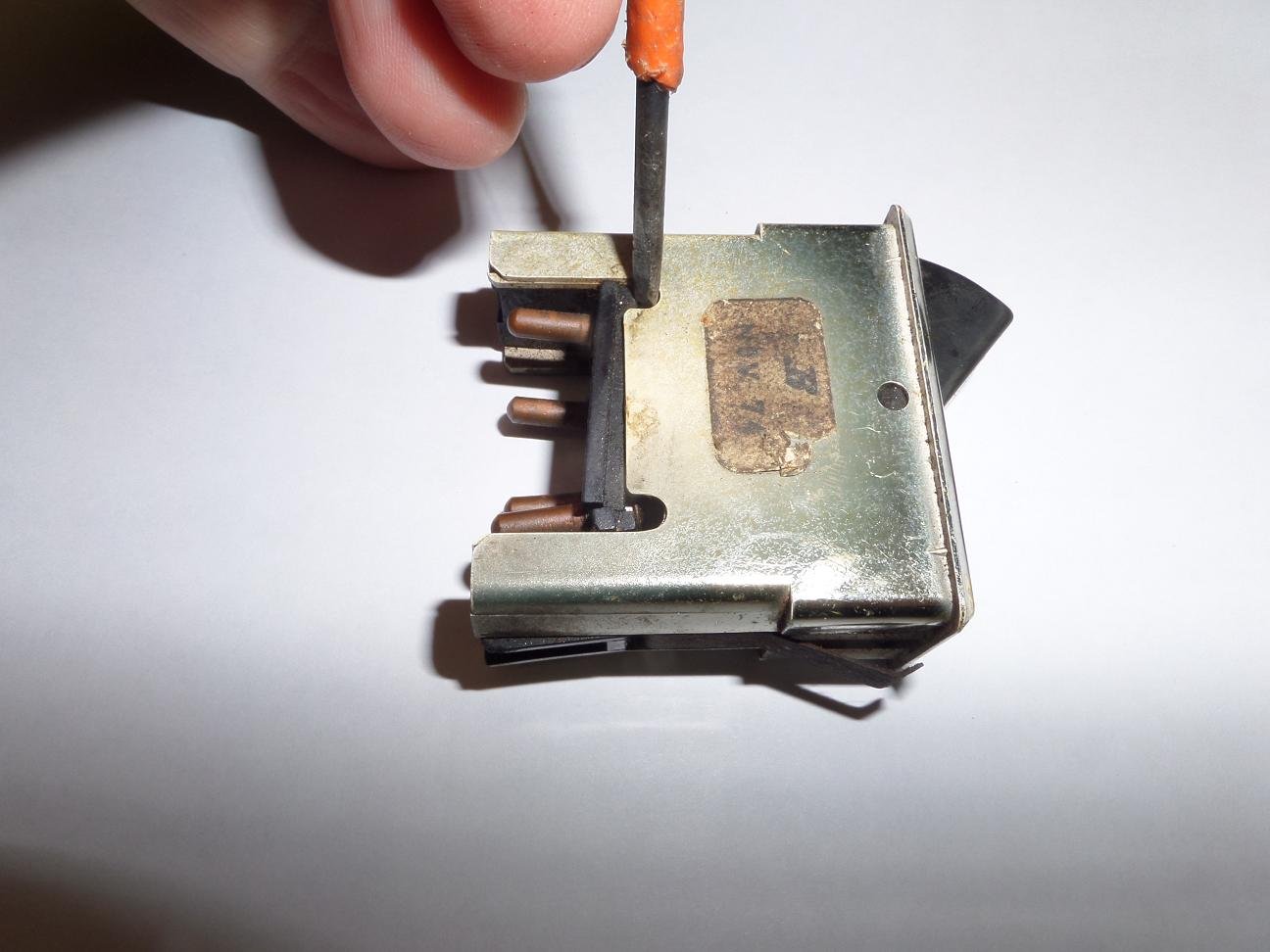 Close-up of a metallic mechanical component with small copper parts and a person's hand holding an orange-handled screwdriver, against a white background.