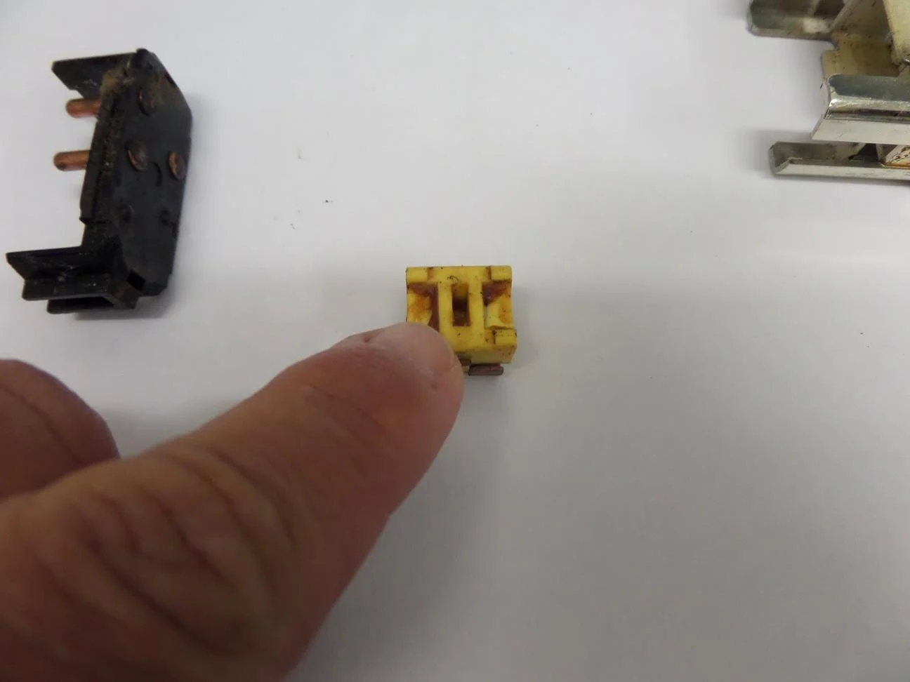 A person's finger holding a small yellow electrical component with rusted contacts, placed on a white surface with other electrical parts nearby.