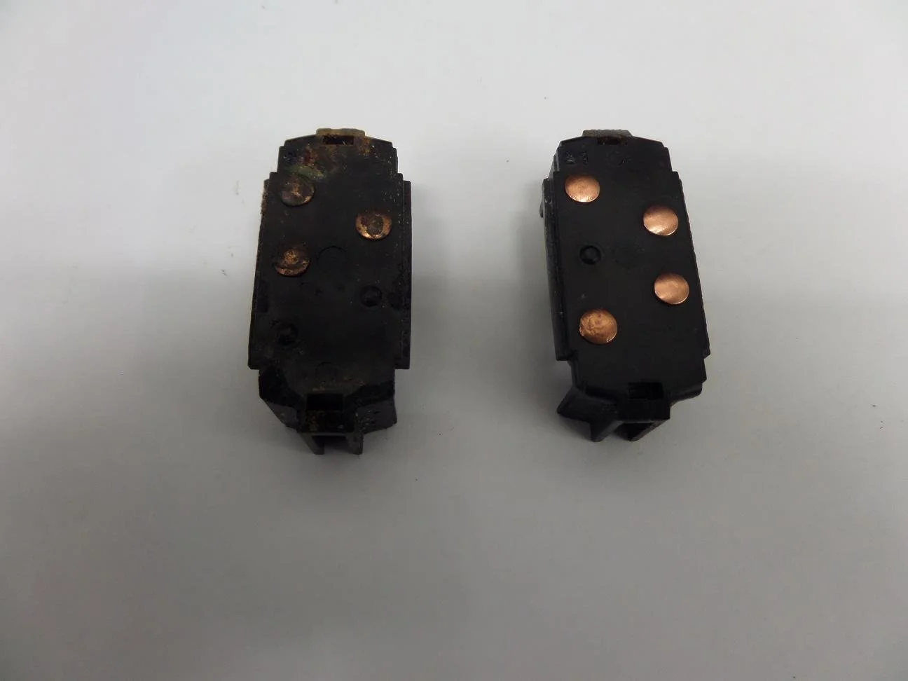 Two black mechanical parts with copper contact points, one with rust and corrosion, placed on a white surface.
