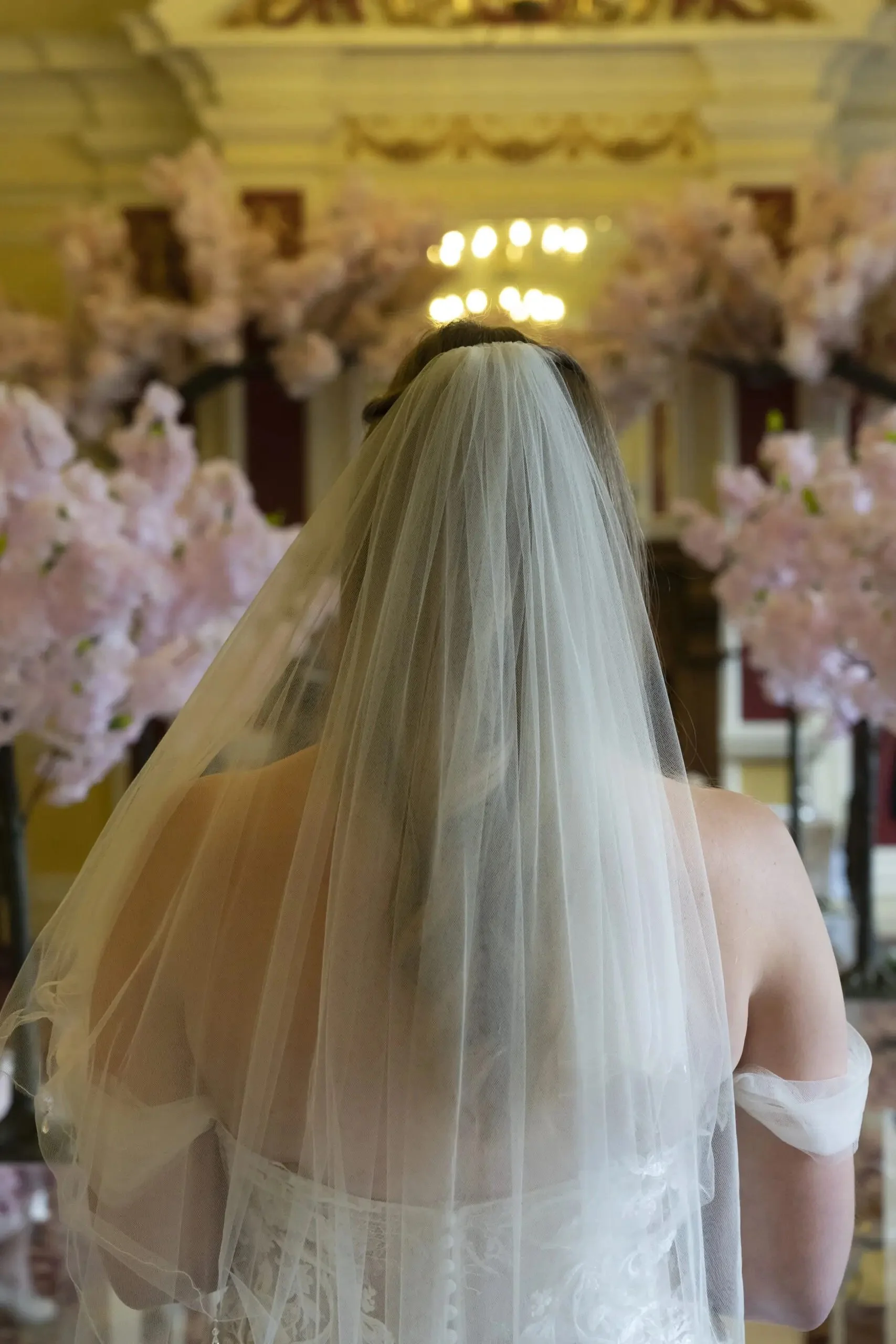 Back view of a bride with a veil, standing in a decorated venue with pink floral arrangements and ornate decor.