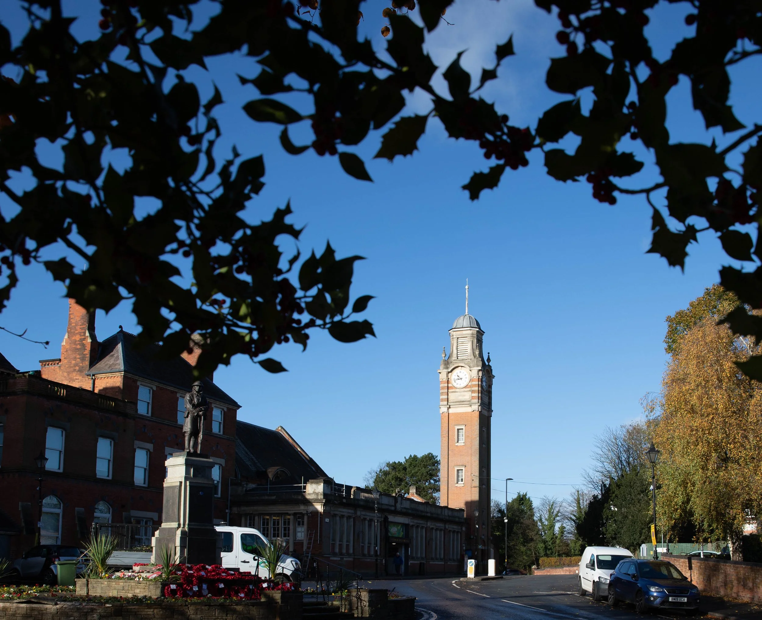 A clock tower with a spire is seen in a small town square, with trees on the right and a statue in the foreground. The sky is clear and blue.