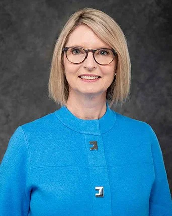 A woman with blond hair and glasses smiling, wearing a bright blue blazer, against a dark gray background.