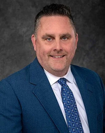 Professional headshot of a man in a blue suit and patterned tie, smiling against a dark gray background.