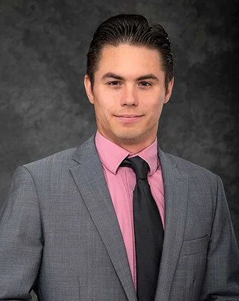 A young man in a light gray suit, pink shirt, and black tie posing against a dark textured background.