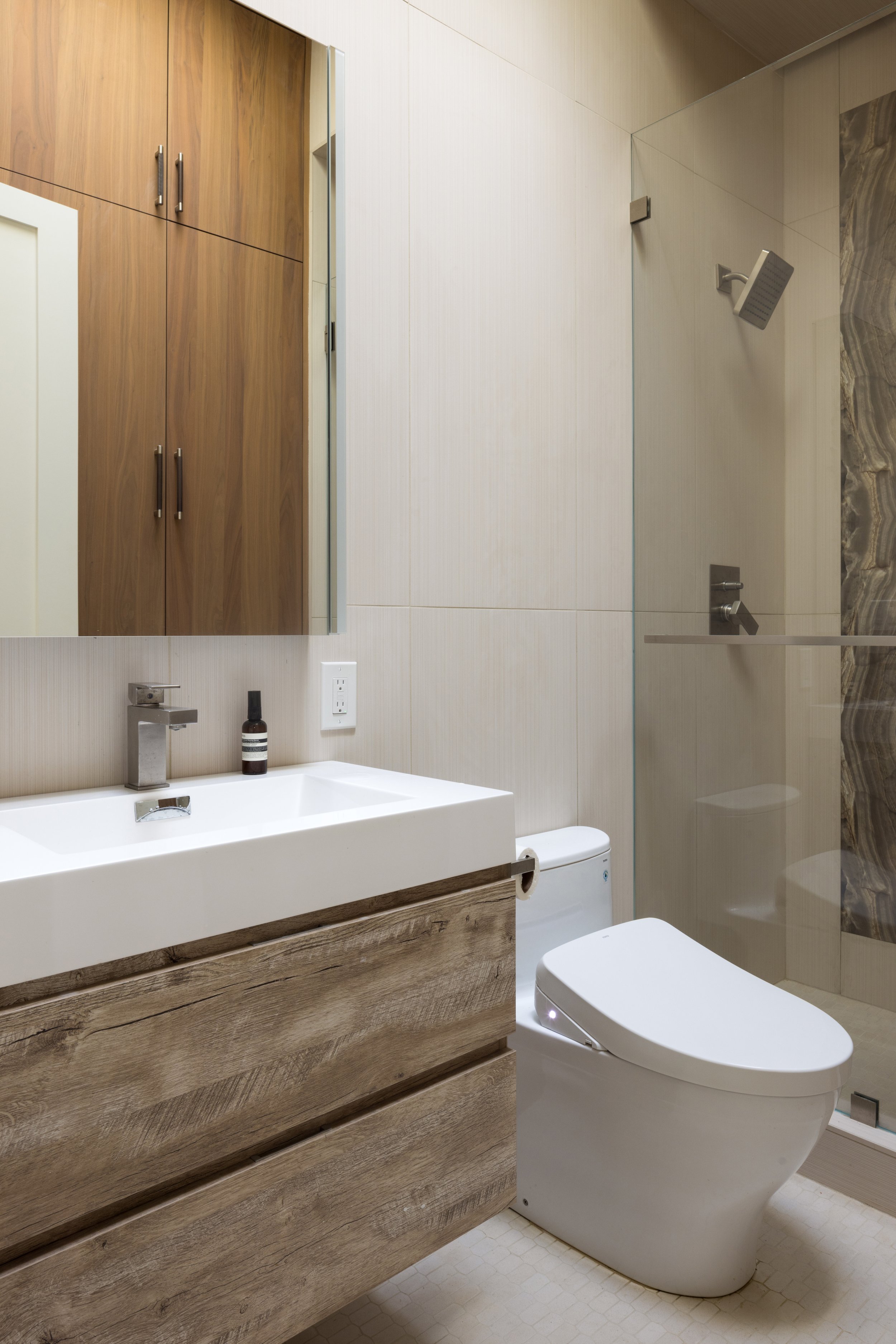 This contemporary bathroom renovation combines large-format porcelain tile, custom walnut cabinetry, and a striking bookmatched onyx feature wall to create a refined spa-like experience. Frameless glass shower enclosures, integrated lighting, and cle