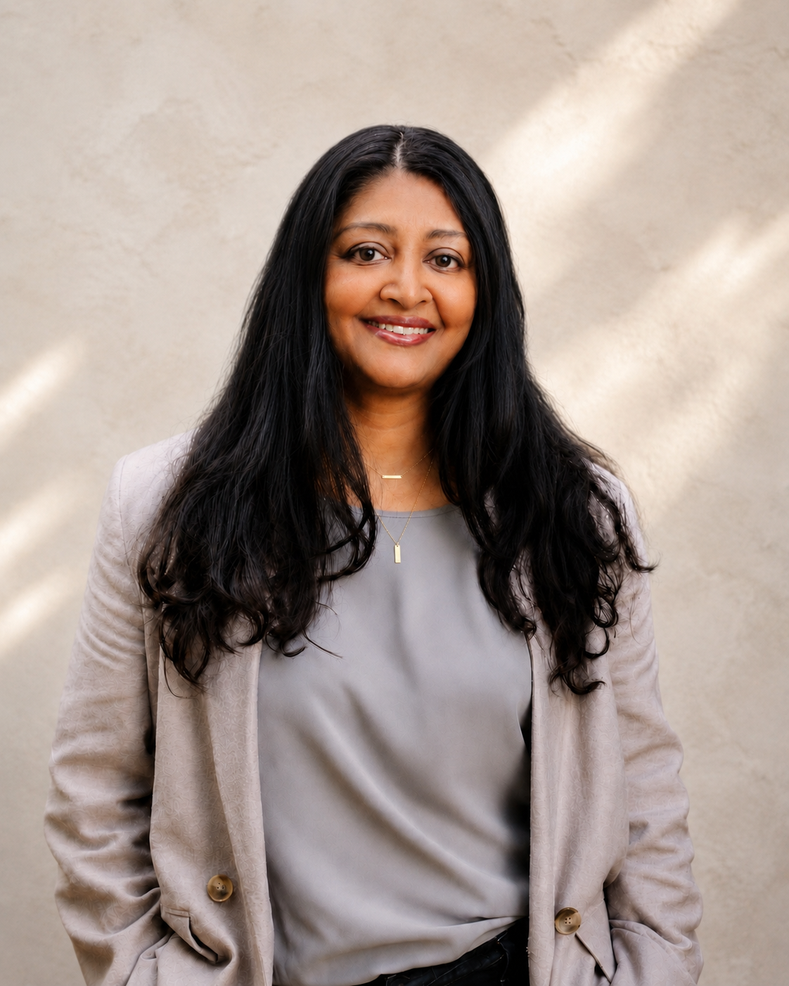 A woman with long black hair, smiling, wearing a beige blazer over a gray blouse, with a necklace, standing against a neutral background.