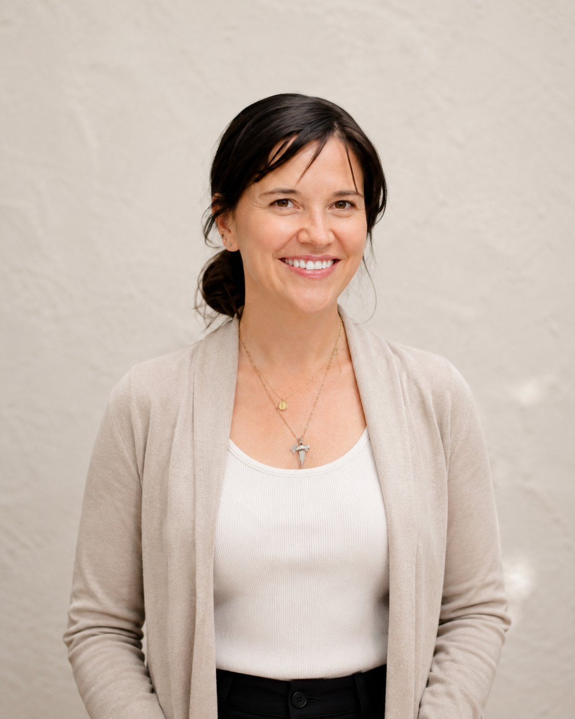 A woman with dark hair tied back, wearing a light beige blazer, white top, black pants, and layered necklaces, smiling and standing against a plain off-white wall.