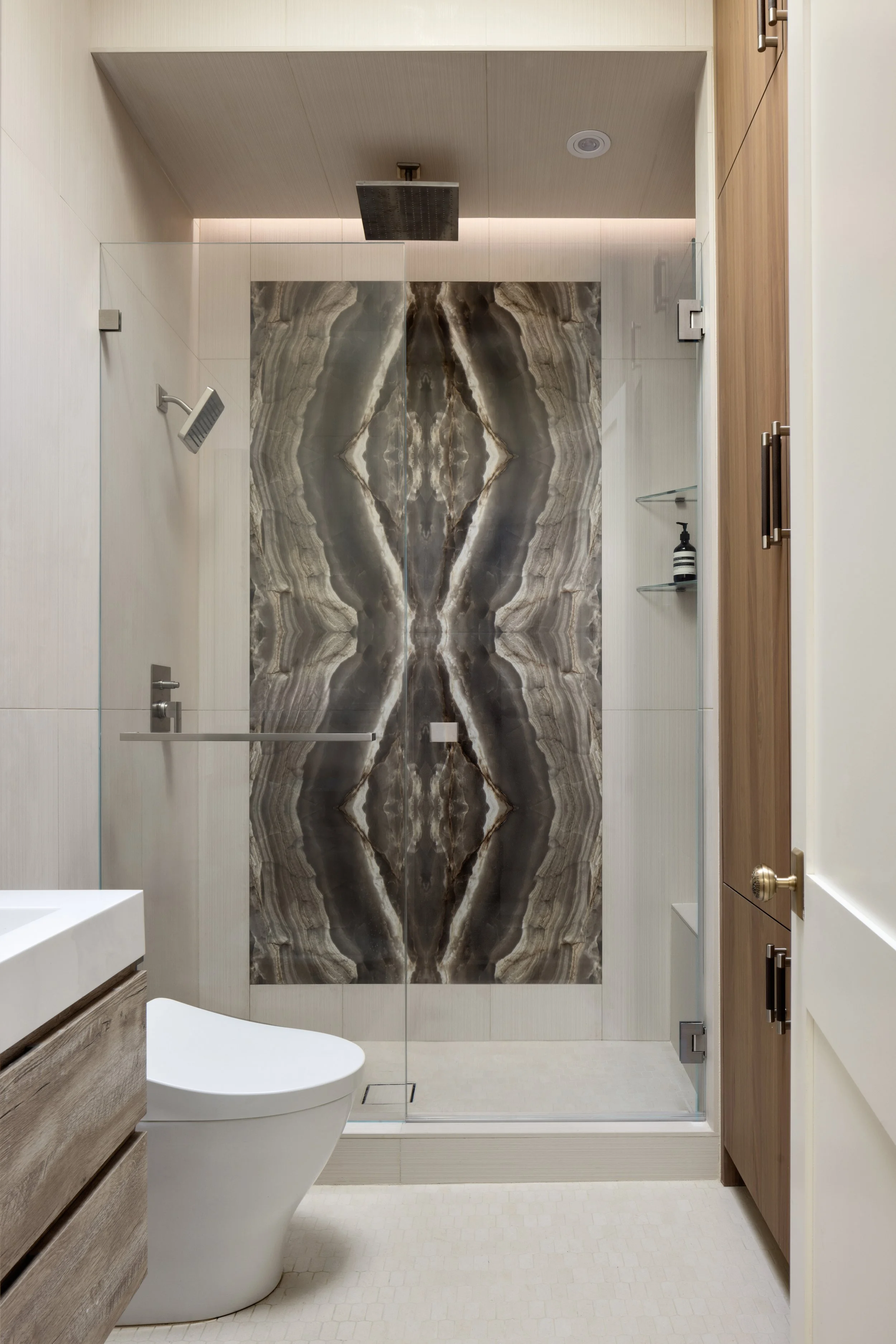 This contemporary bathroom renovation combines large-format porcelain tile, custom walnut cabinetry, and a striking bookmatched onyx feature wall to create a refined spa-like experience. Frameless glass shower enclosures, integrated lighting, and cle