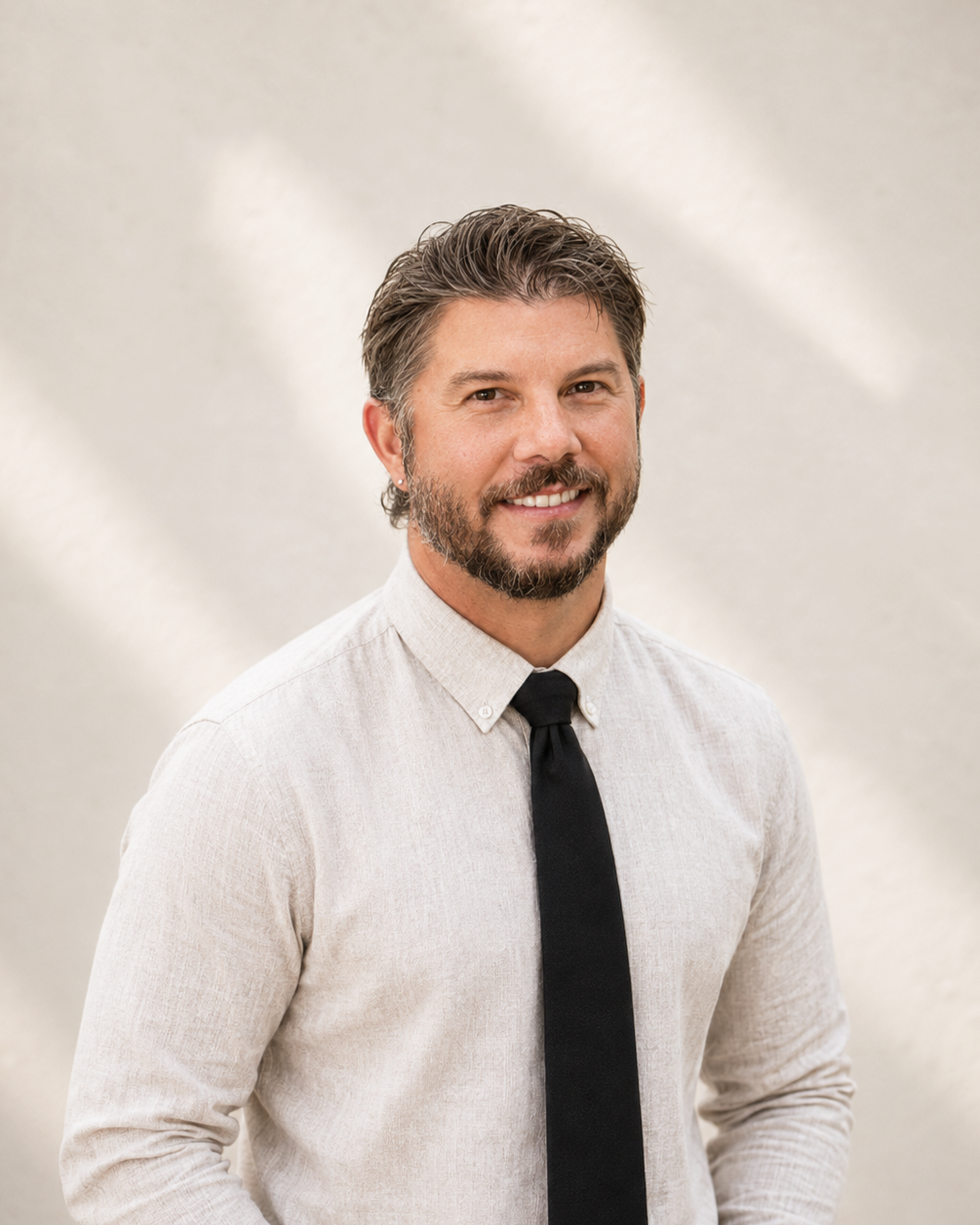 A smiling man with dark blonde hair, a beard, and a mustache wearing a light beige shirt and a black tie, standing against a neutral background.