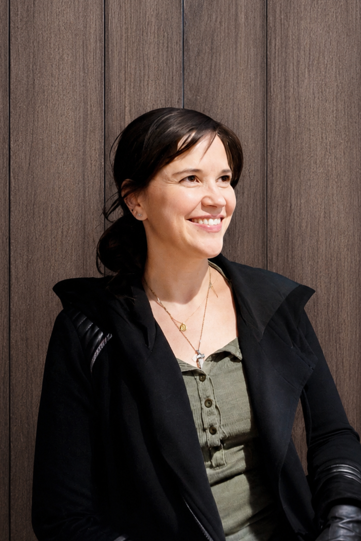 Smiling person with dark hair wearing a black jacket and light-colored top in front of a wooden background.