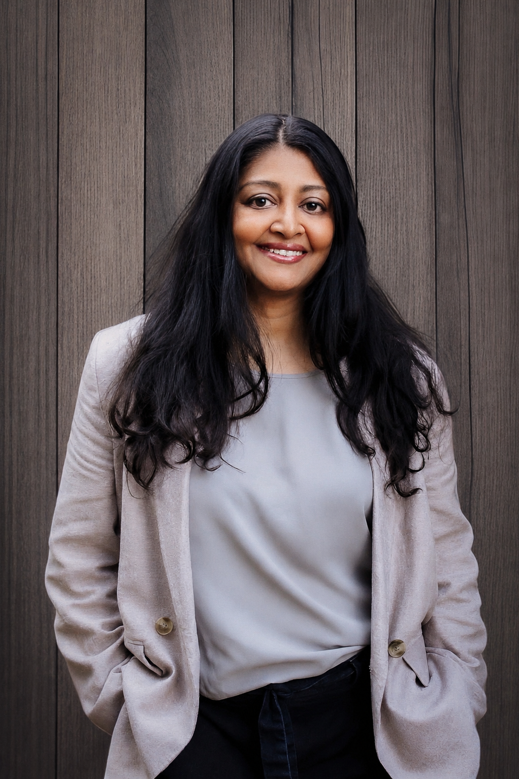 Person with long dark hair wearing a light blazer and gray top against a wooden background.