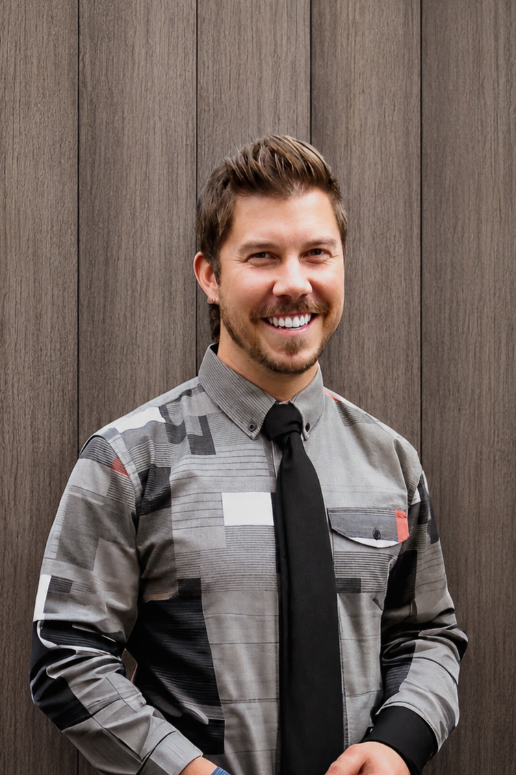 Man wearing a patterned shirt and tie, standing in front of a wood panel background.