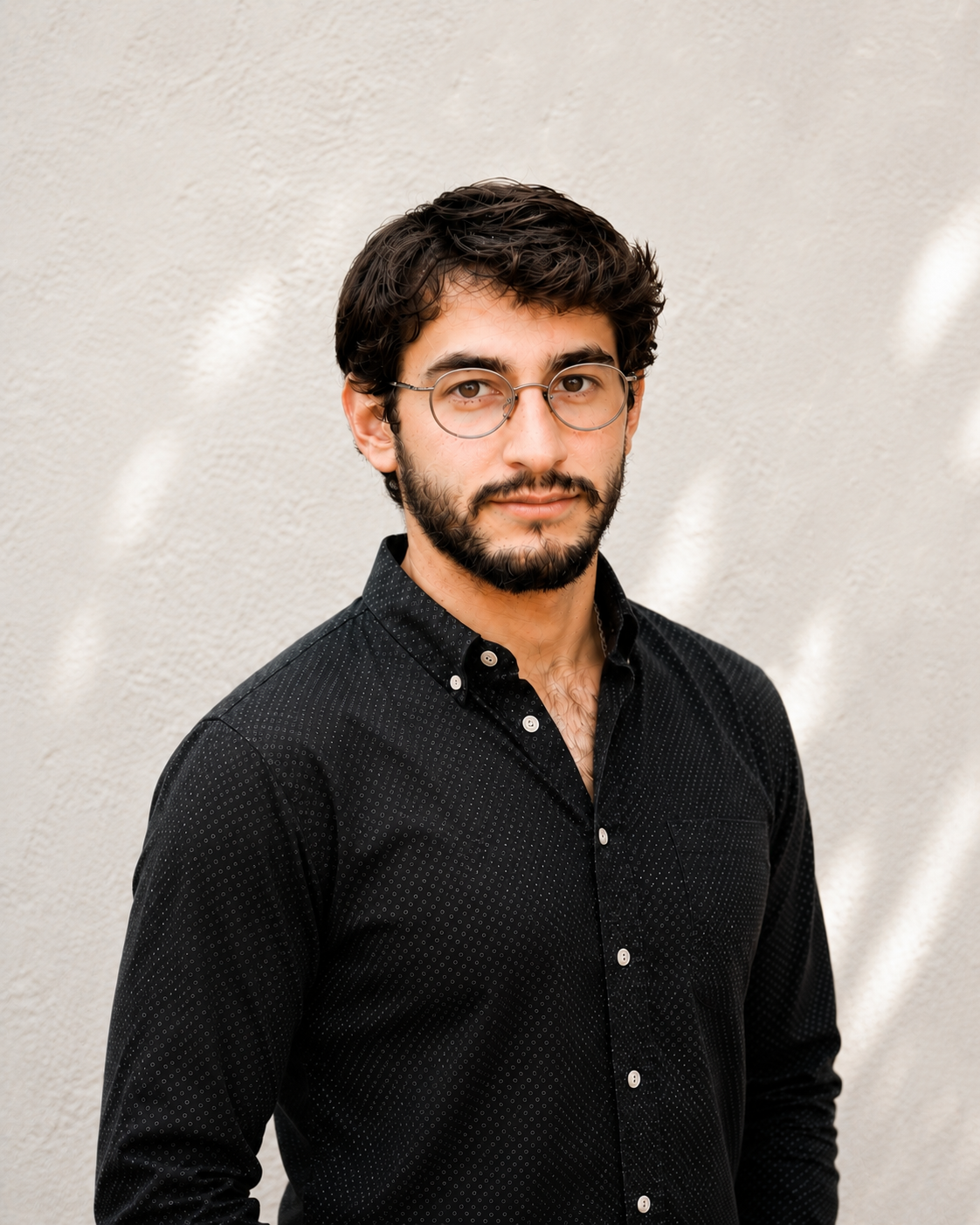 A man with dark hair, glasses, and a beard wearing a black button-up shirt standing against a light textured background.