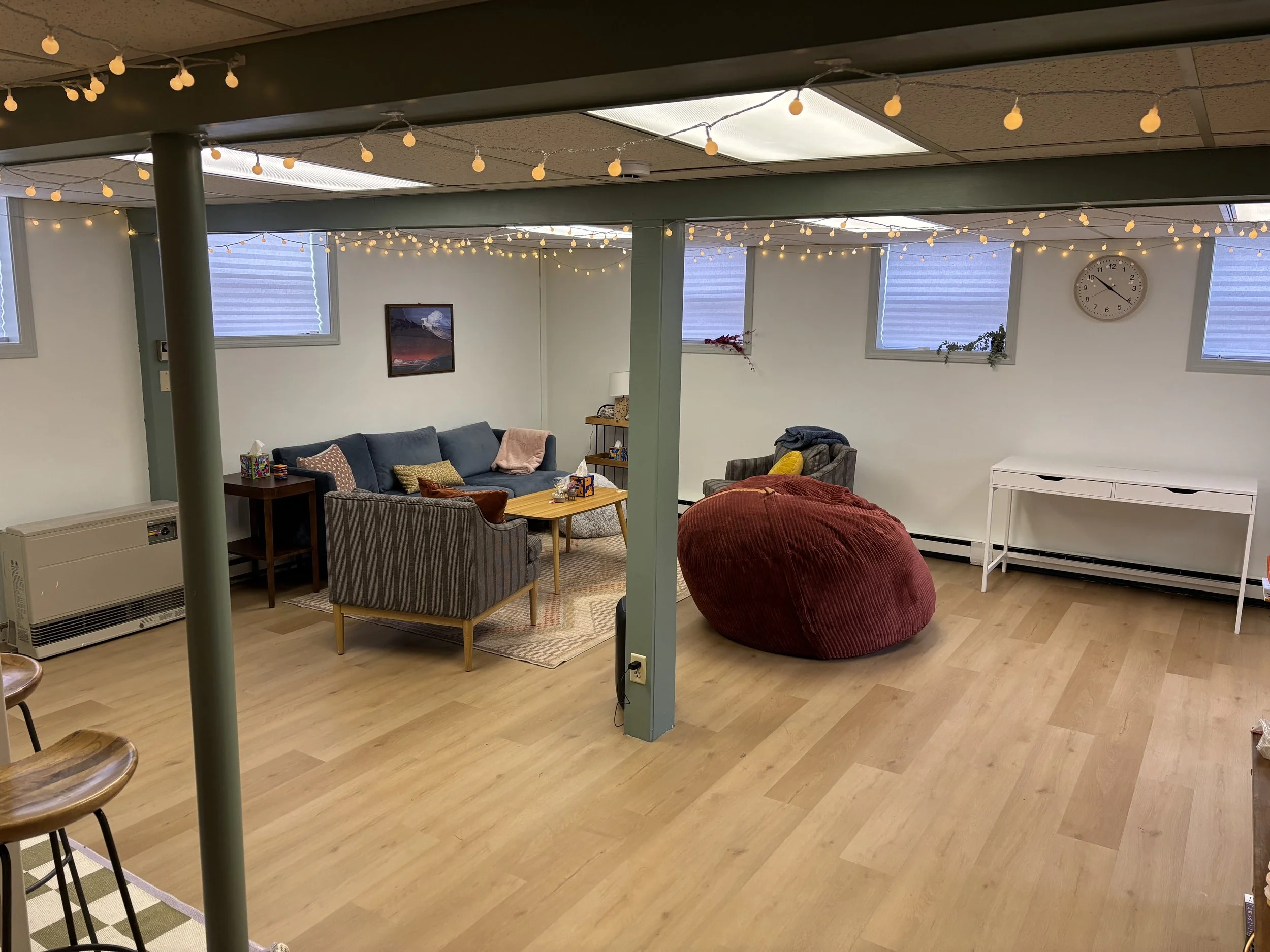 Spacious basement living room with string lights, a navy sofa, striped armchair, red bean bag, wall clock, windows with blinds, and a white table.