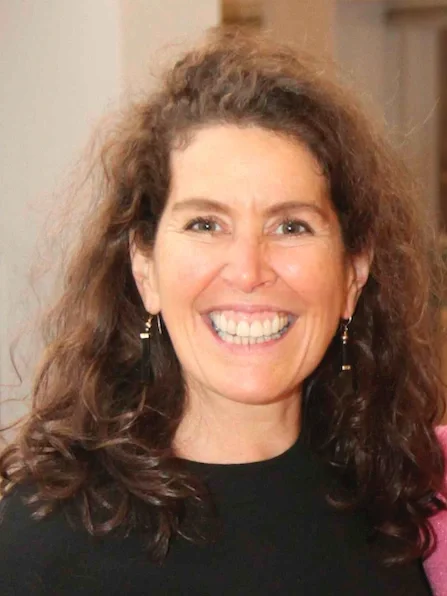 A smiling woman with curly brown hair, wearing earrings and a black top, indoors.