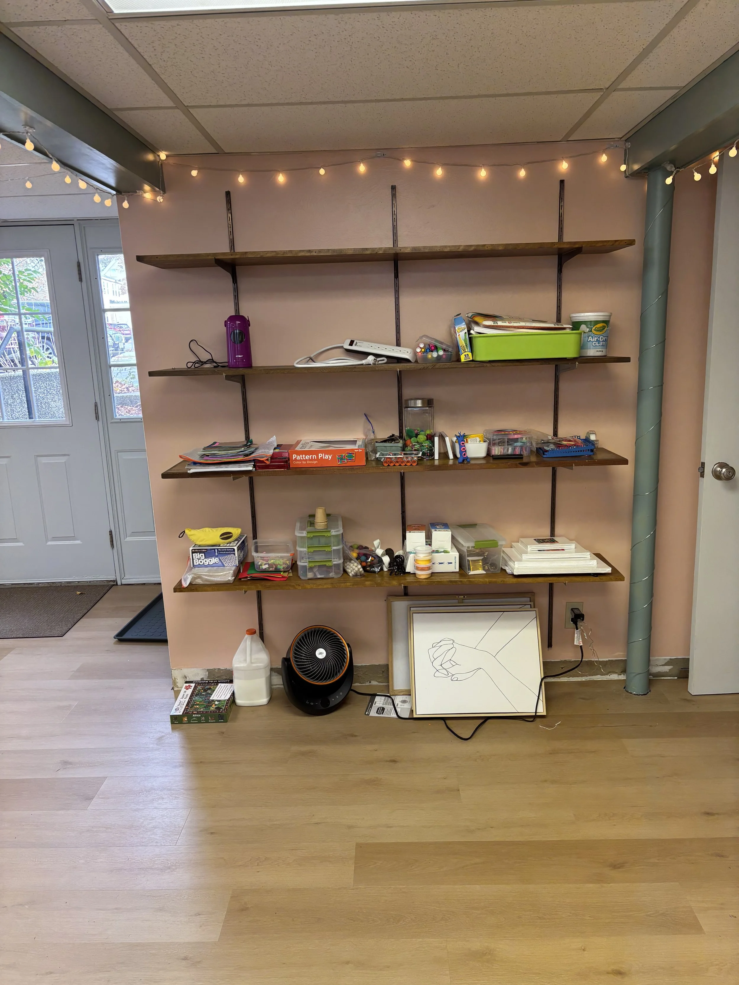 A corner of a room with a pink wall and wooden floor, featuring a wall-mounted wooden shelf with miscellaneous items and a string of small yellow lights along the ceiling. On the floor below the shelves, there are various objects including a whiteboa