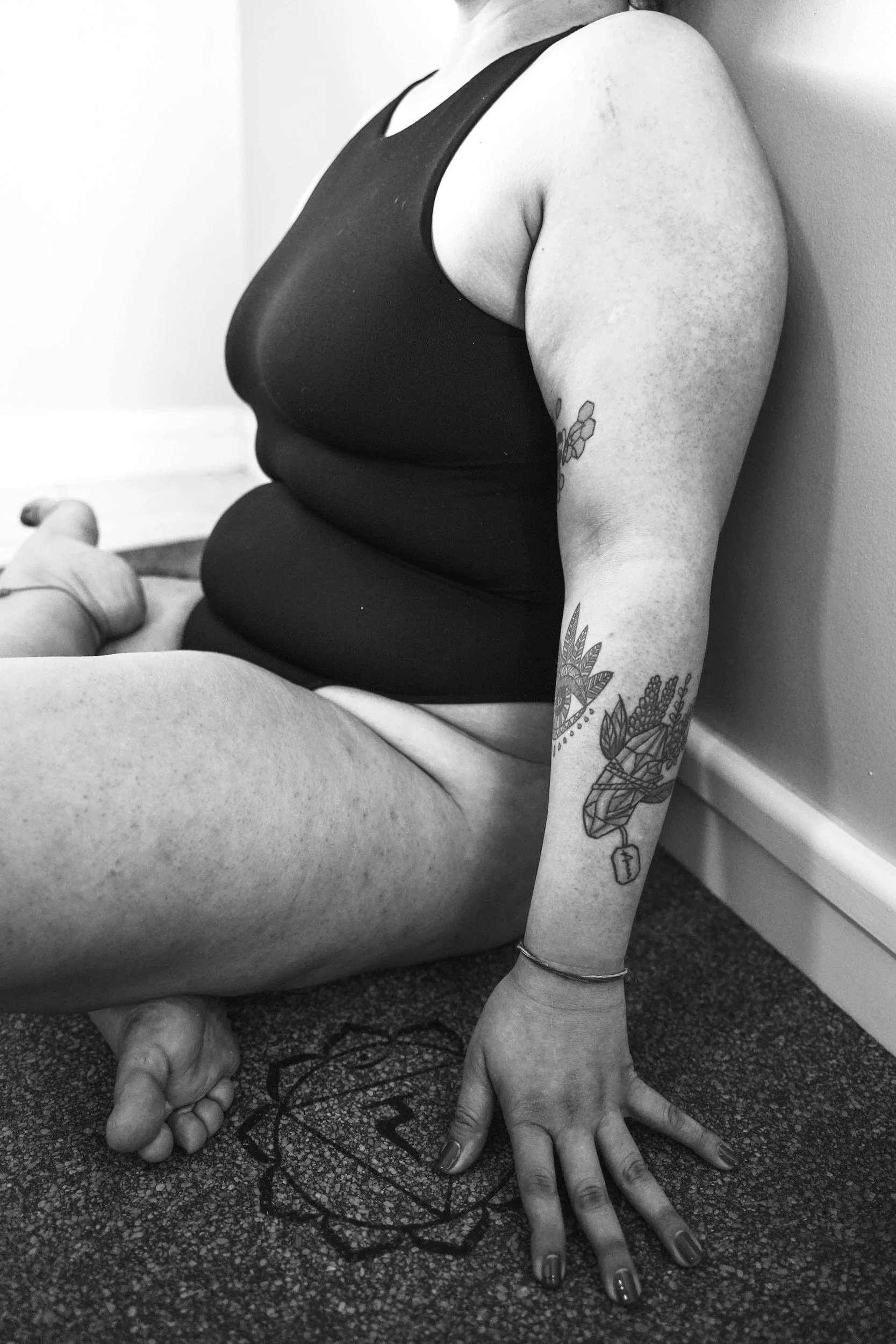 A woman sitting on the floor with her back against the wall, wearing a tank top. Her arm with tattoos rests on the floor, and her hand is near a mandala and a Peace sign on the carpet. She has a tattoo of a geometric fox and plants on her arm.