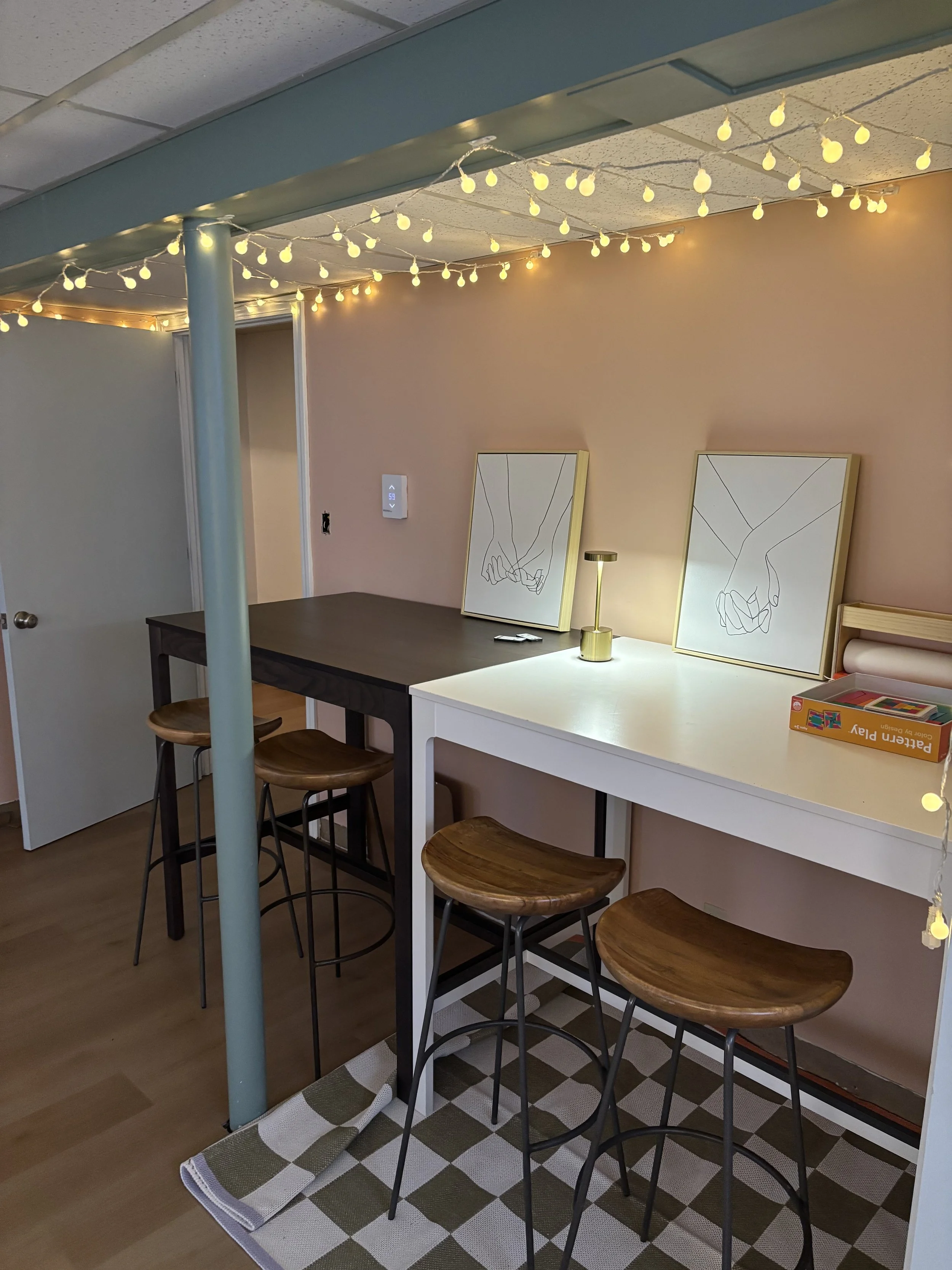 Cozy indoor space with string lights, a small bar-height table with three wooden stools, two framed line art prints of holding hands on the wall, a small gold lamp, and a board game box on a white countertop.
