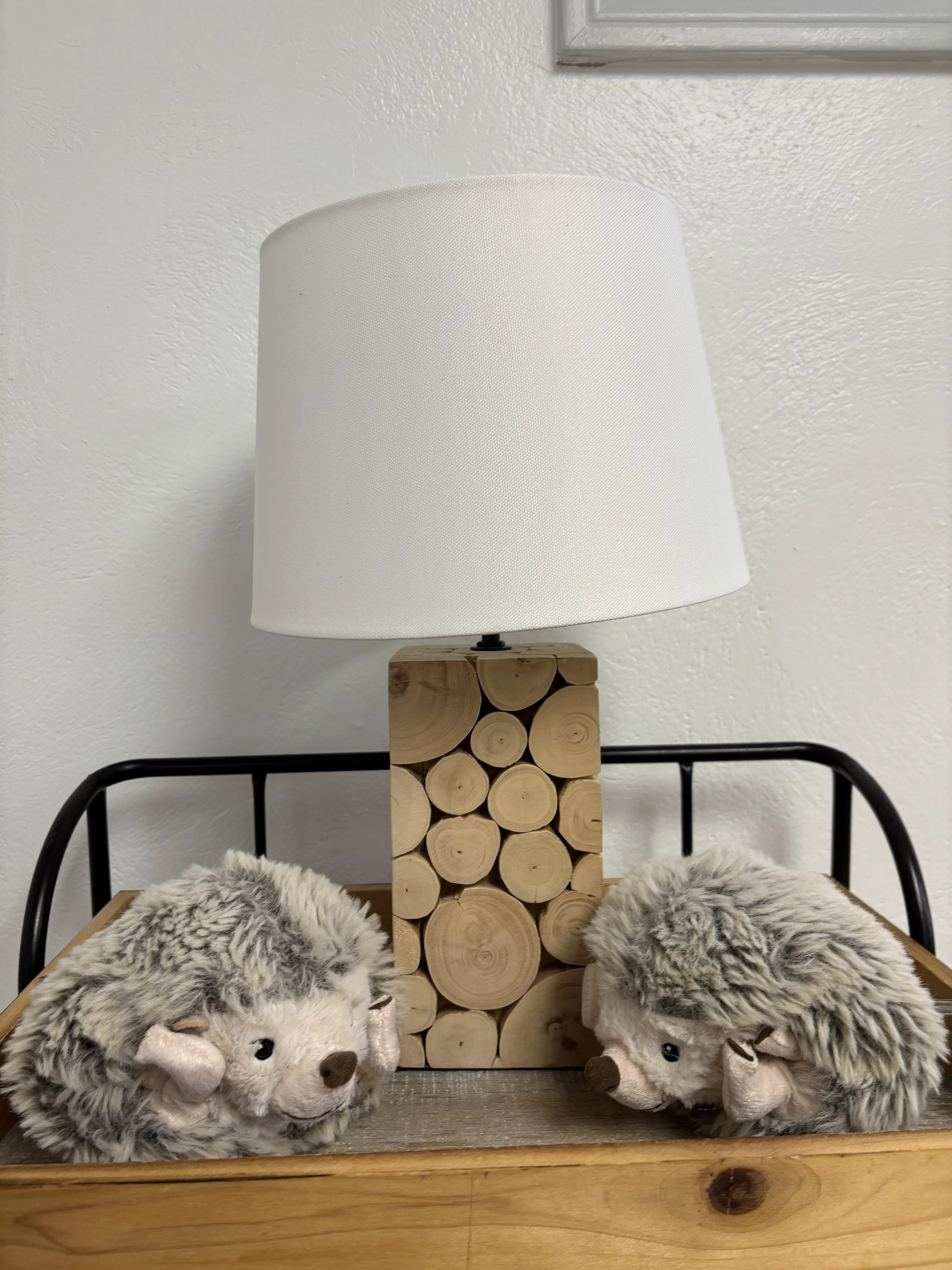 Decorative table lamp with a white shade, situated on a wooden tray with two plush hedgehog toys and a stack of wooden logs in the background.