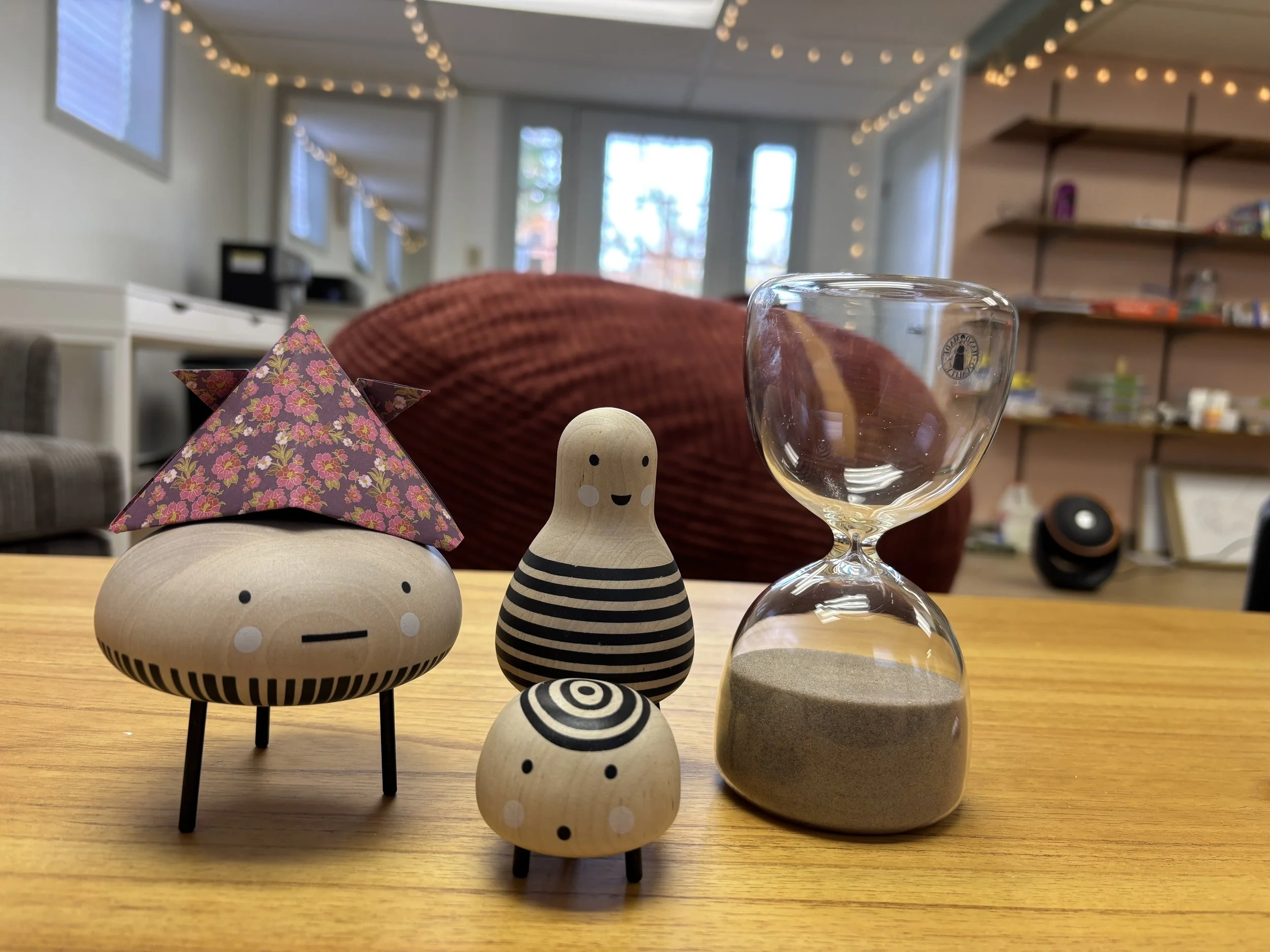 Decorative wooden toys with faces, a pyramid-shaped item wrapped in pink floral paper, an hourglass with sand, set on a wooden table in a cozy room with string lights and a window in the background.