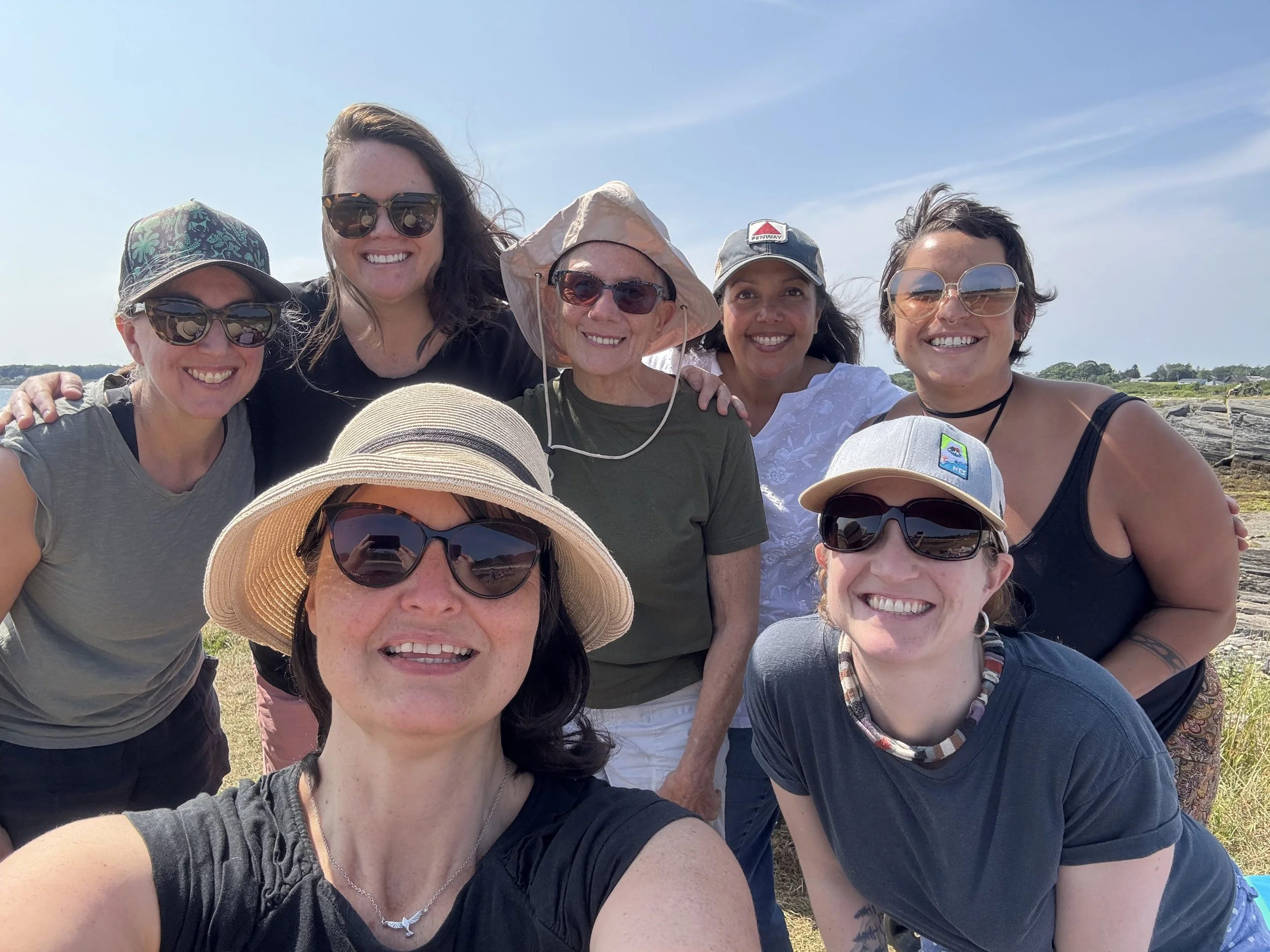 Group of eight smiling women outdoors on a sunny day, some wearing sunglasses and hats, with a rocky landscape and blue sky in the background.
