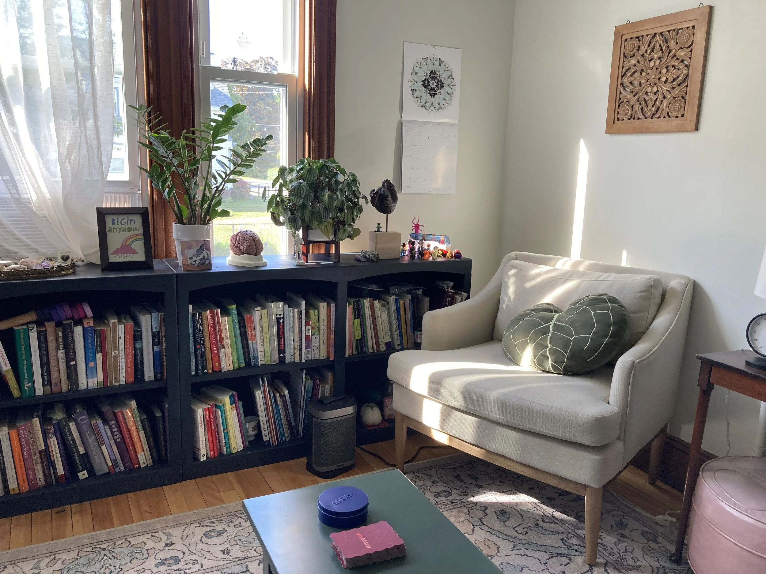 Living room with a window, bookshelf, potted plants, decorative items, a white armchair with a green pillow, a side table with a clock, and a patterned rug.