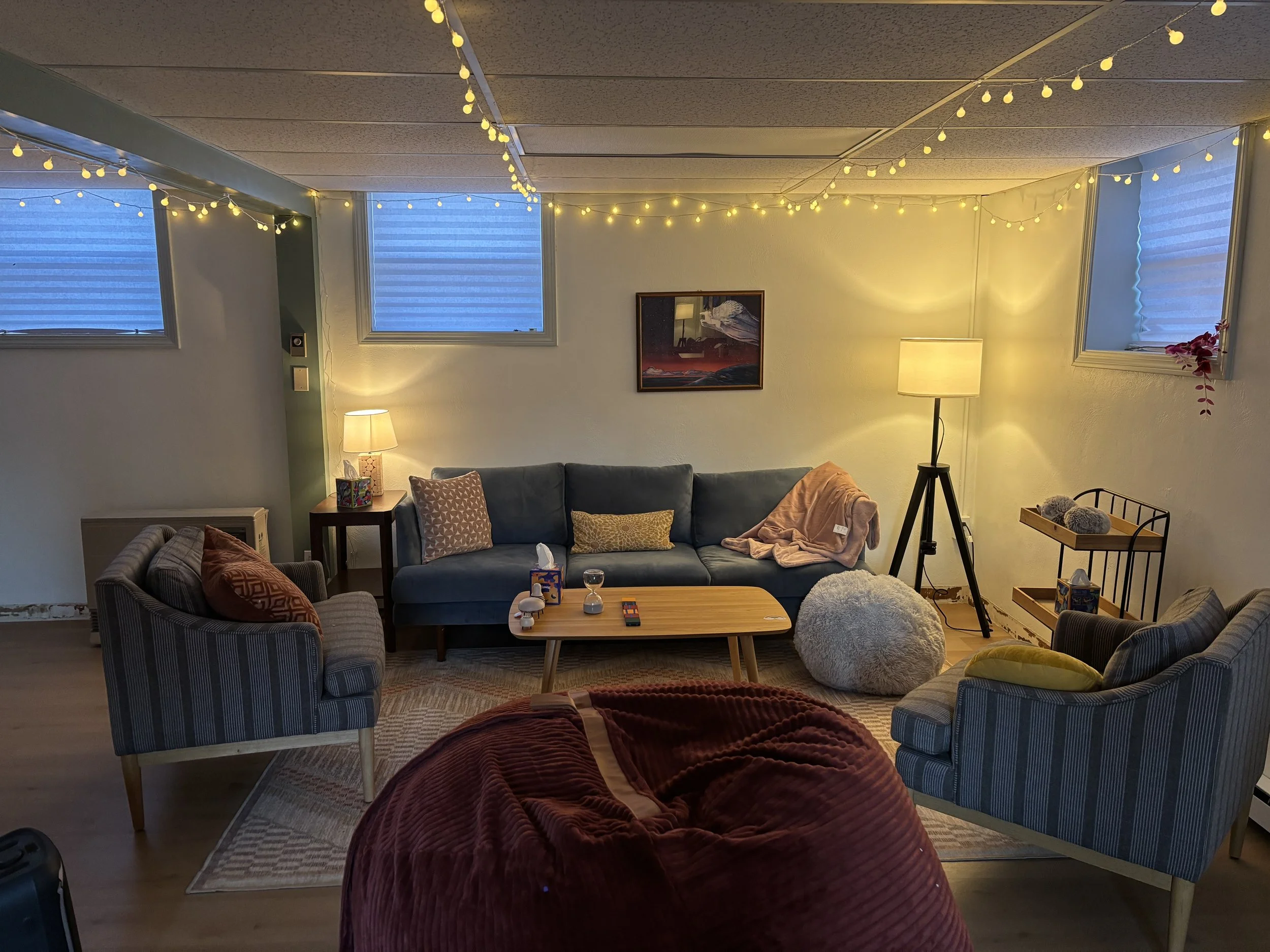 Cozy living room with blue couch, patterned armchairs, string lights, multiple lamps, framed artwork, small wooden coffee table, plush ottovar, and decorative pillows.
