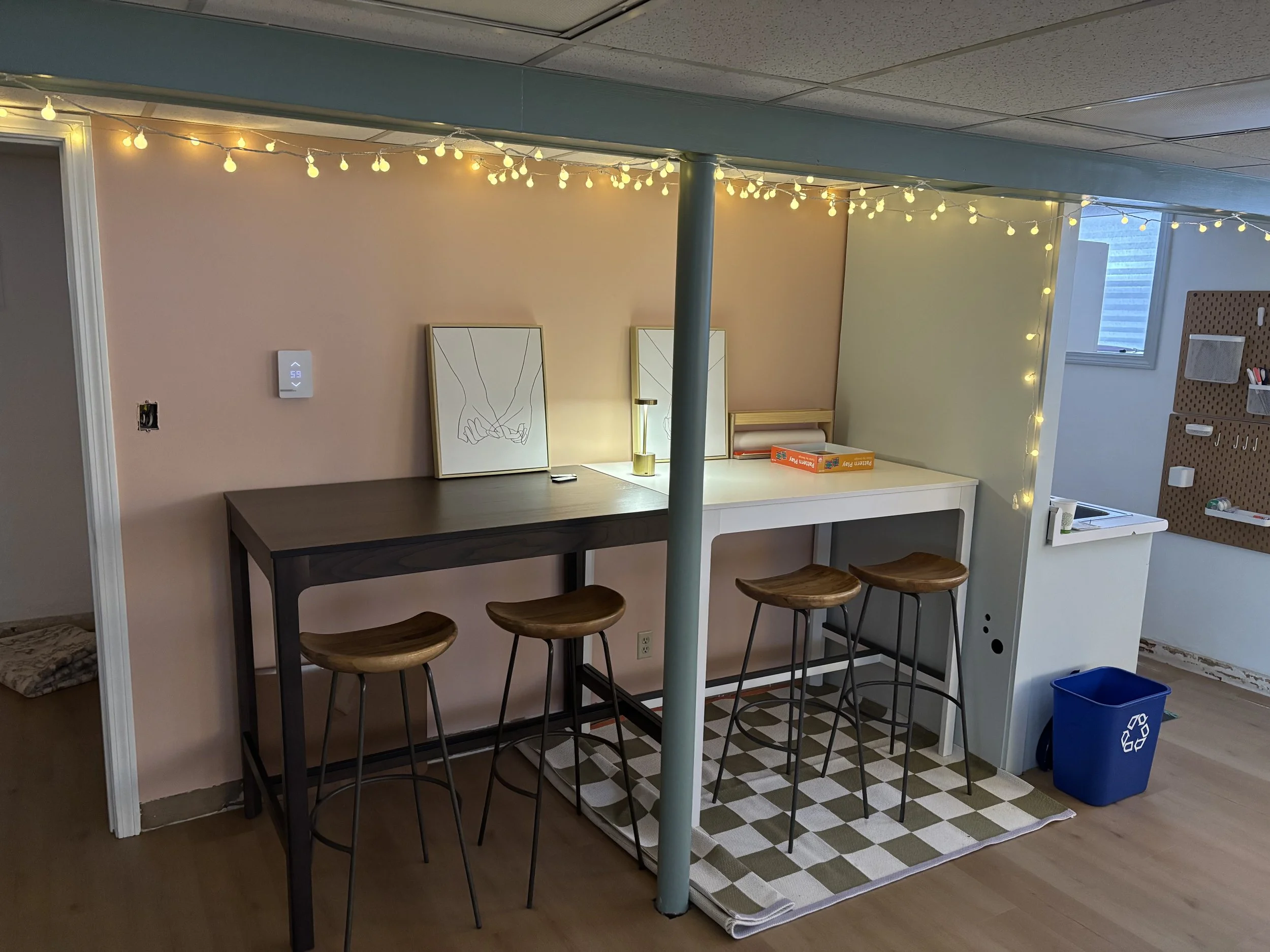 A cozy room corner with string lights, a pink wall, two framed line art prints, a black high table with three wooden stools, a white desk with a small lamp, a box of Jenga, a pegboard with supplies, and a recycling bin.
