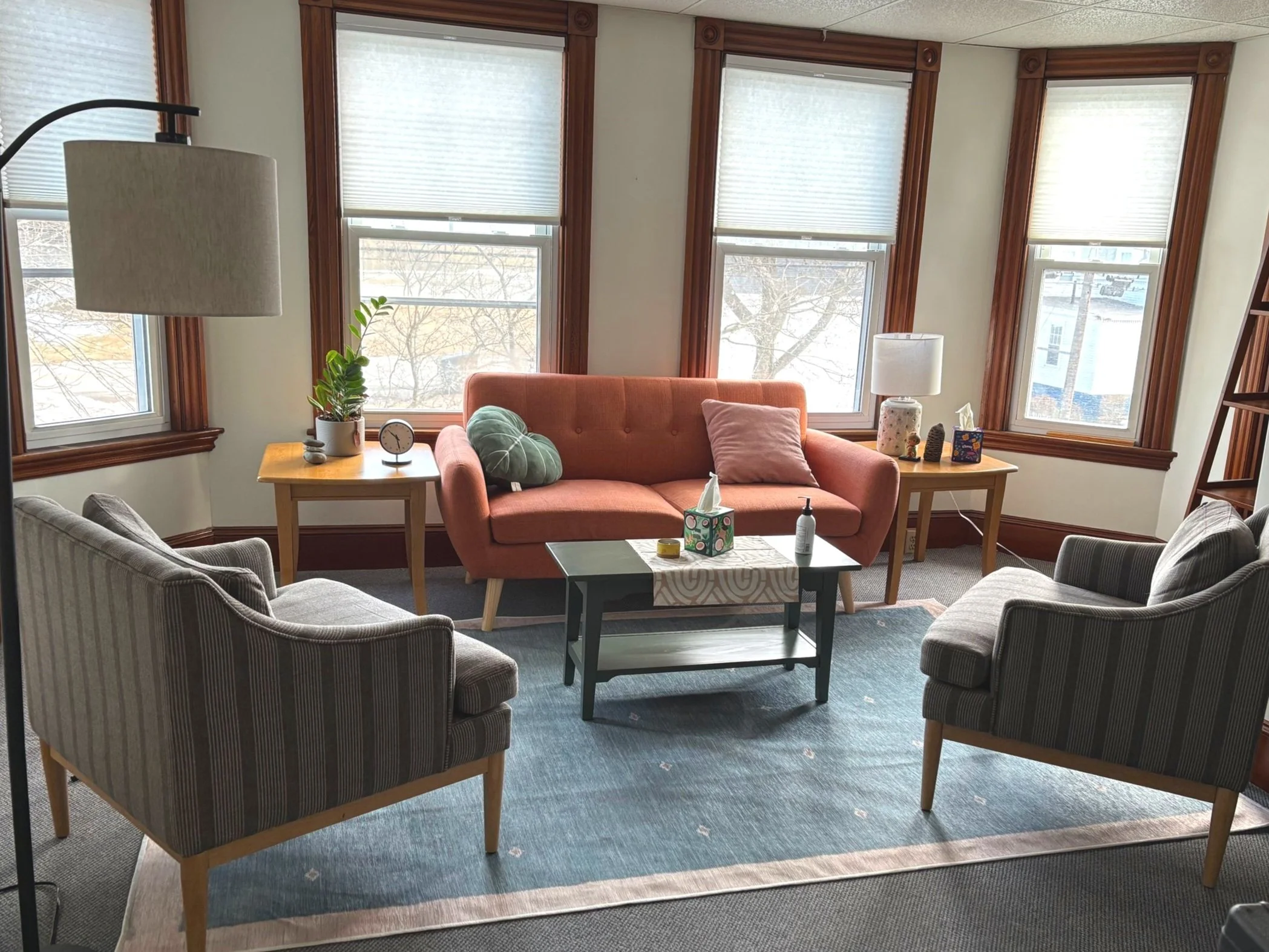 Living room with four striped armchairs, a peach couch, a light blue rug, three windows with wooden trim, a wooden side table with a plant and clock, a black coffee table with tissue box and hand sanitizer, and side tables with lamps and decorative items.