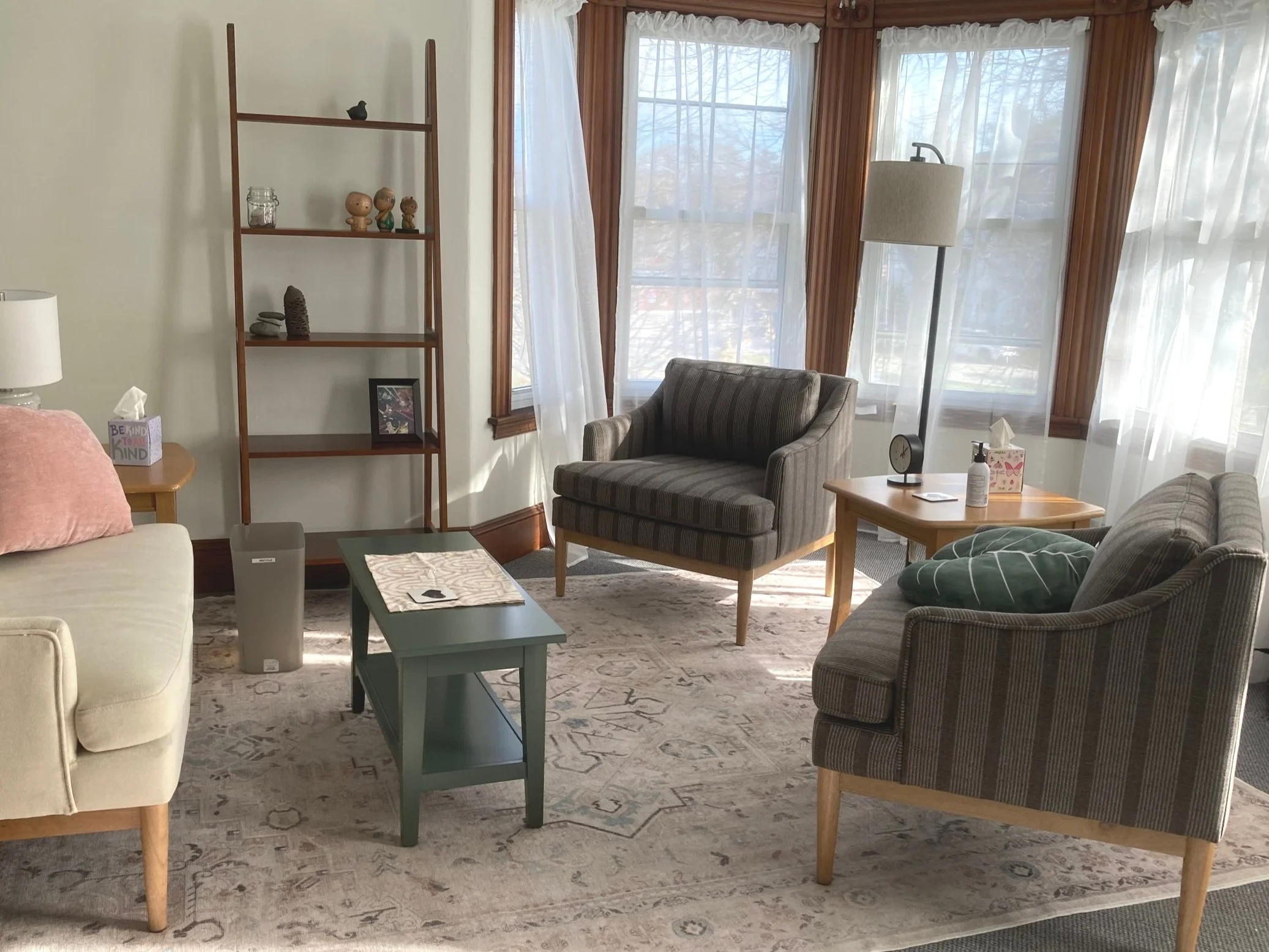 Cozy living room with beige, striped, and gray armchairs, a small green table, a standing floor lamp, and a wooden side table with tissues, a clock, and sanitizer. Large windows with sheer curtains let in natural light, and there is a wooden bookshelf with decorative items and a framed photo.