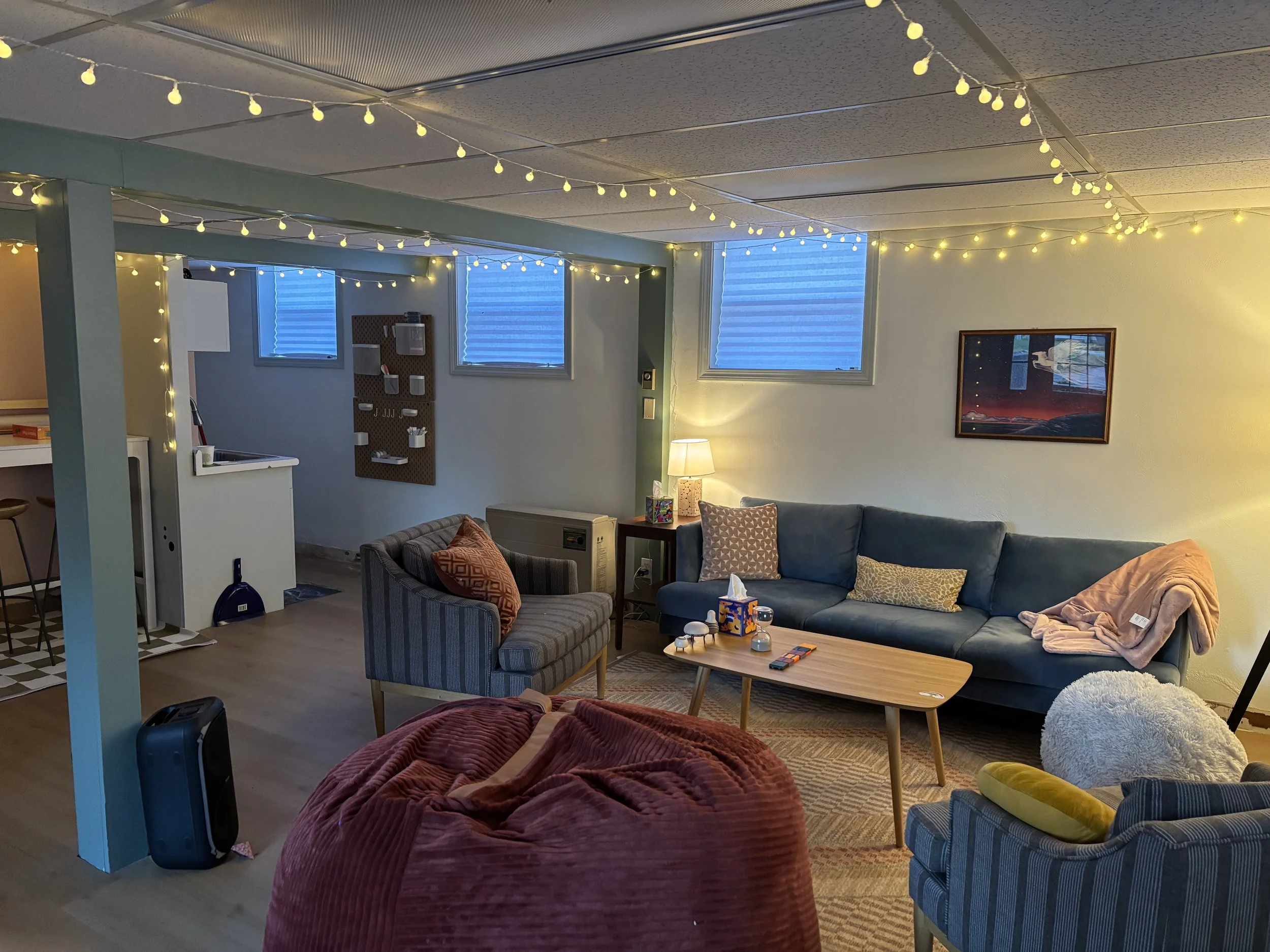 Cozy living room illuminated with string lights, featuring a blue sofa with throw pillows, a wooden coffee table with snacks and decor, a striped armchair, a side table with a lamp, and a wall art. The space has small windows and a warm, inviting atm