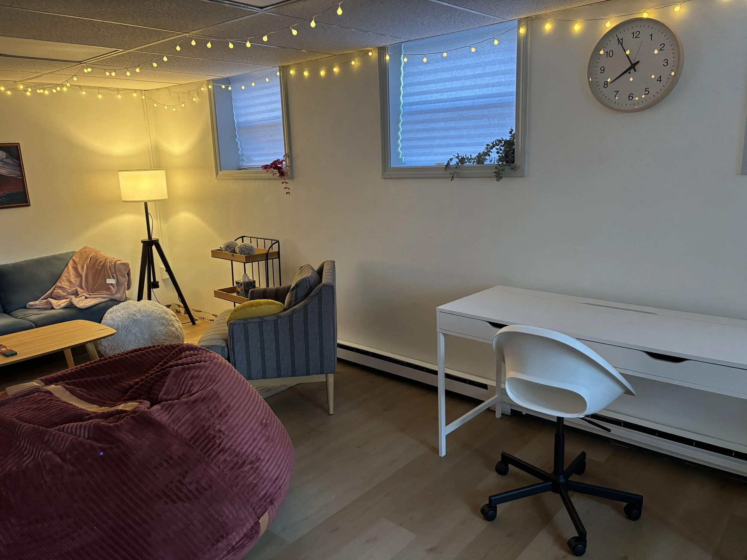 Living room with string lights hanging from the ceiling, a standing lamp illuminating the space, an armchair with a yellow pillow, a sofa with blankets, a white desk with a swivel chair, and a wall clock showing 10:07.