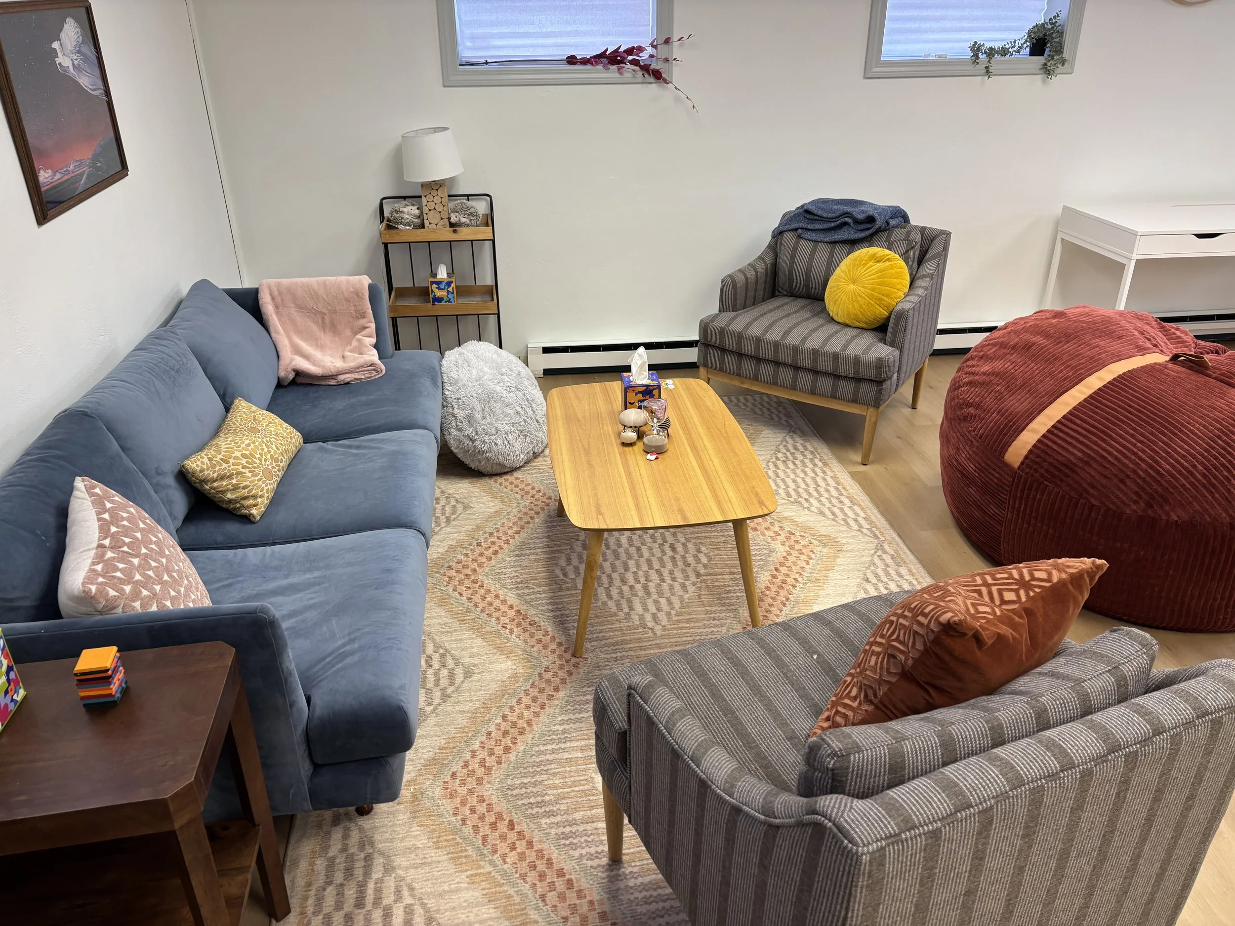 Living room with a blue sofa on the left, a wooden coffee table in the center, a gray armchair with pillows on the right, a red bean bag chair, a small wood side table with a toy on it, a lamp on a shelf, and two windows with plants.