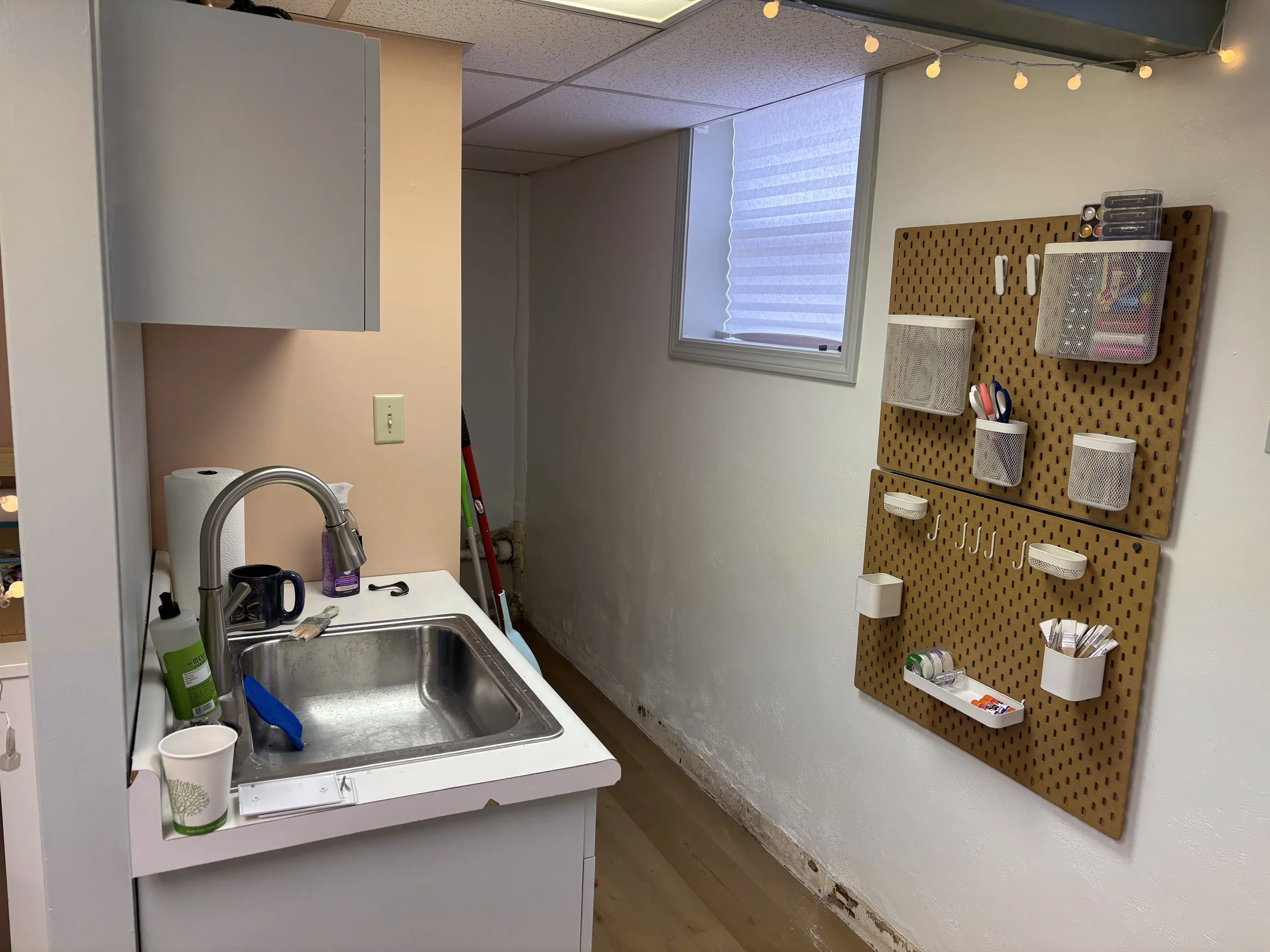 Small kitchen area with a sink, paper towel, mug, and cleaning supplies on the counter. A window with a blind is above the sink. A pegboard with various containers and tools is mounted on the wall to the right.