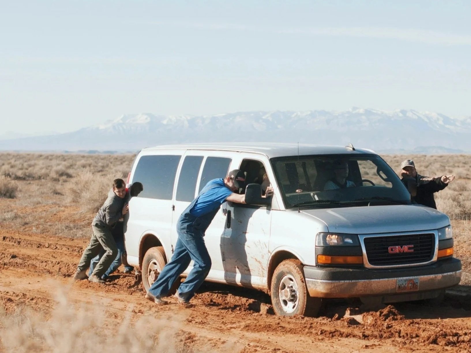 Four people push a white GMC van out of a muddy rut on a dirt road in a barren, open landscape with mountains in the background.