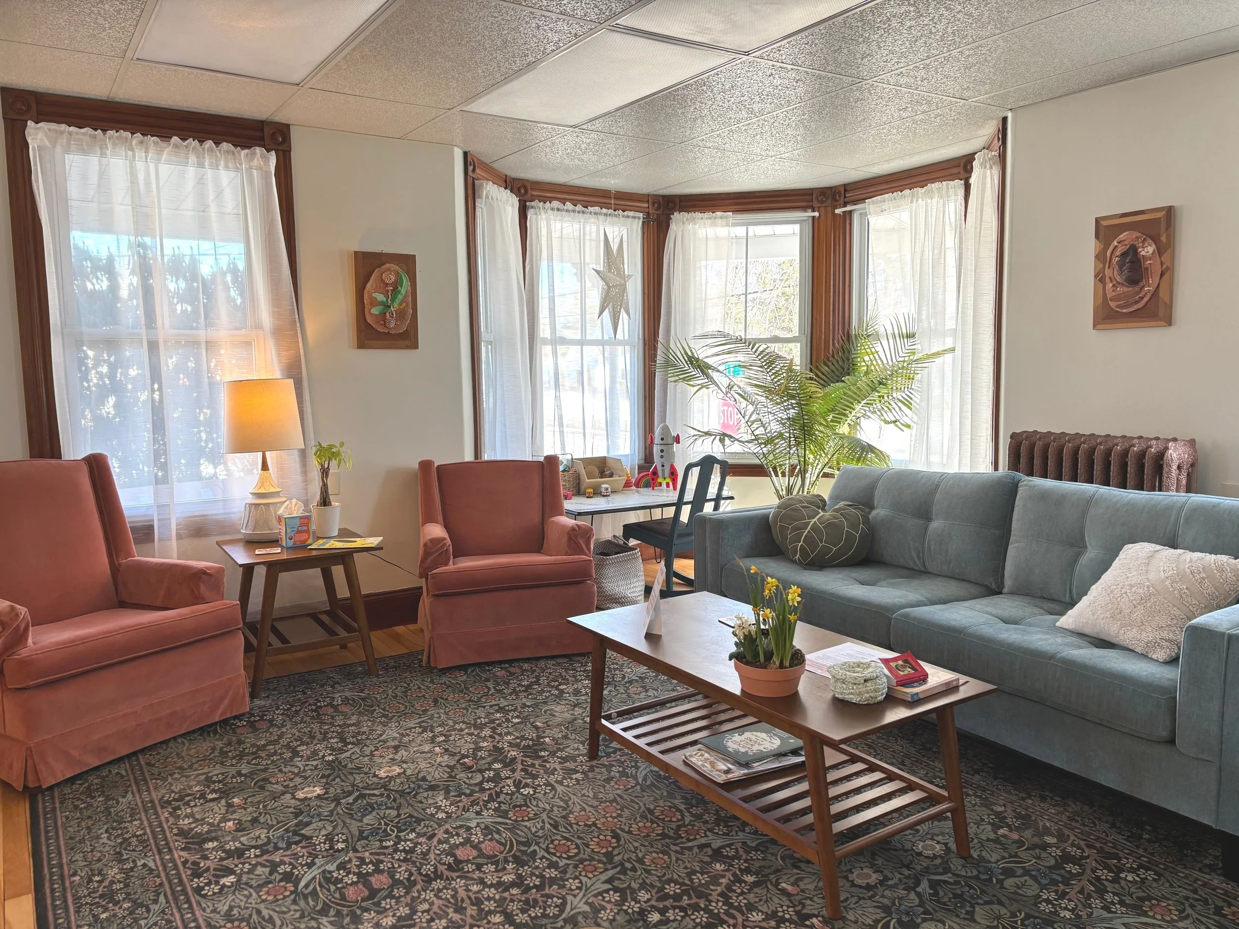 Living room with large window, pink armchairs, a blue sofa, a wooden coffee table with decorations, a side table with a lamp, decorative wall art, and a potted plant.