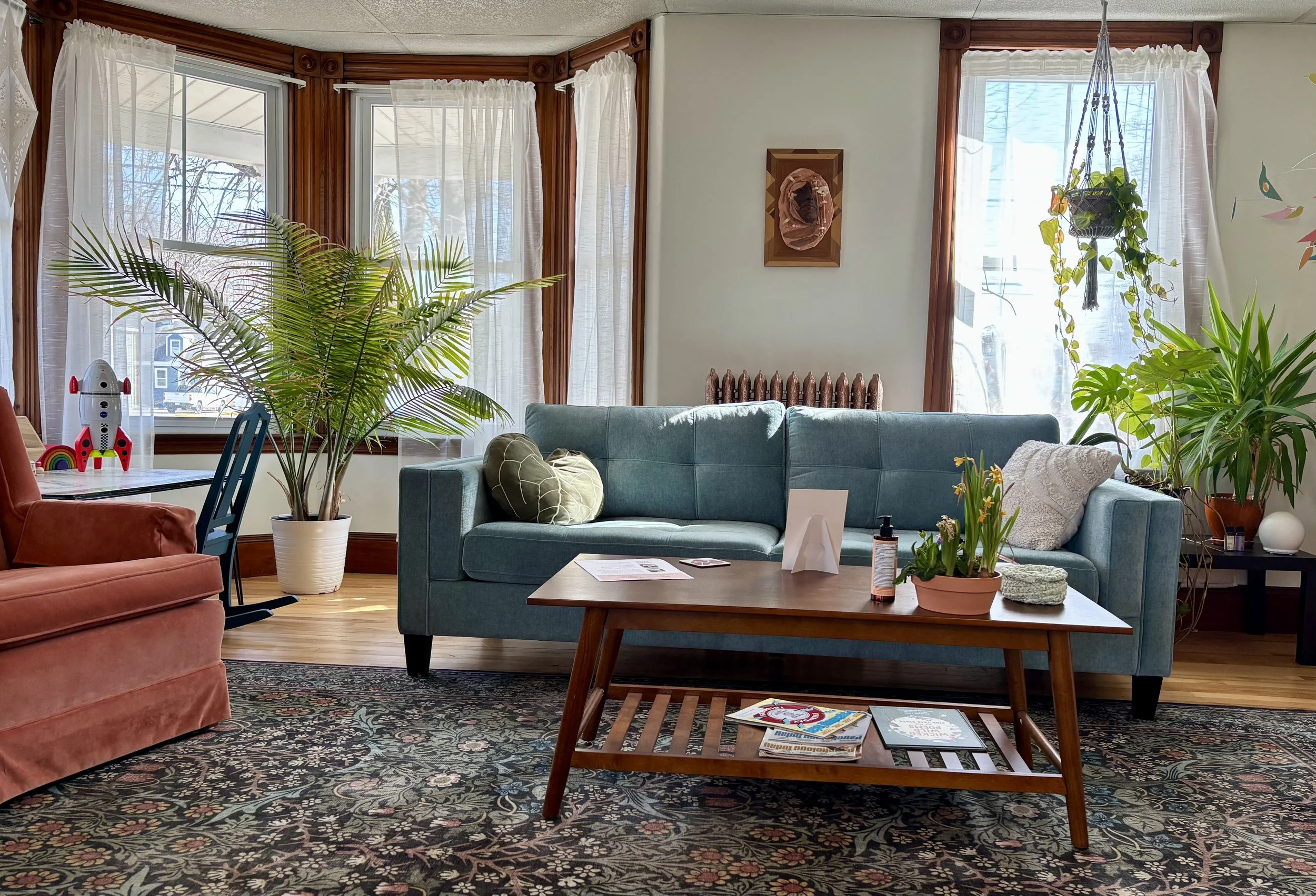 Living room with teal sofa, rust-colored armchair, wooden coffee table, large potted plants, large windows with white curtains, patterned rug, decorative wall art, and various small objects on the table.